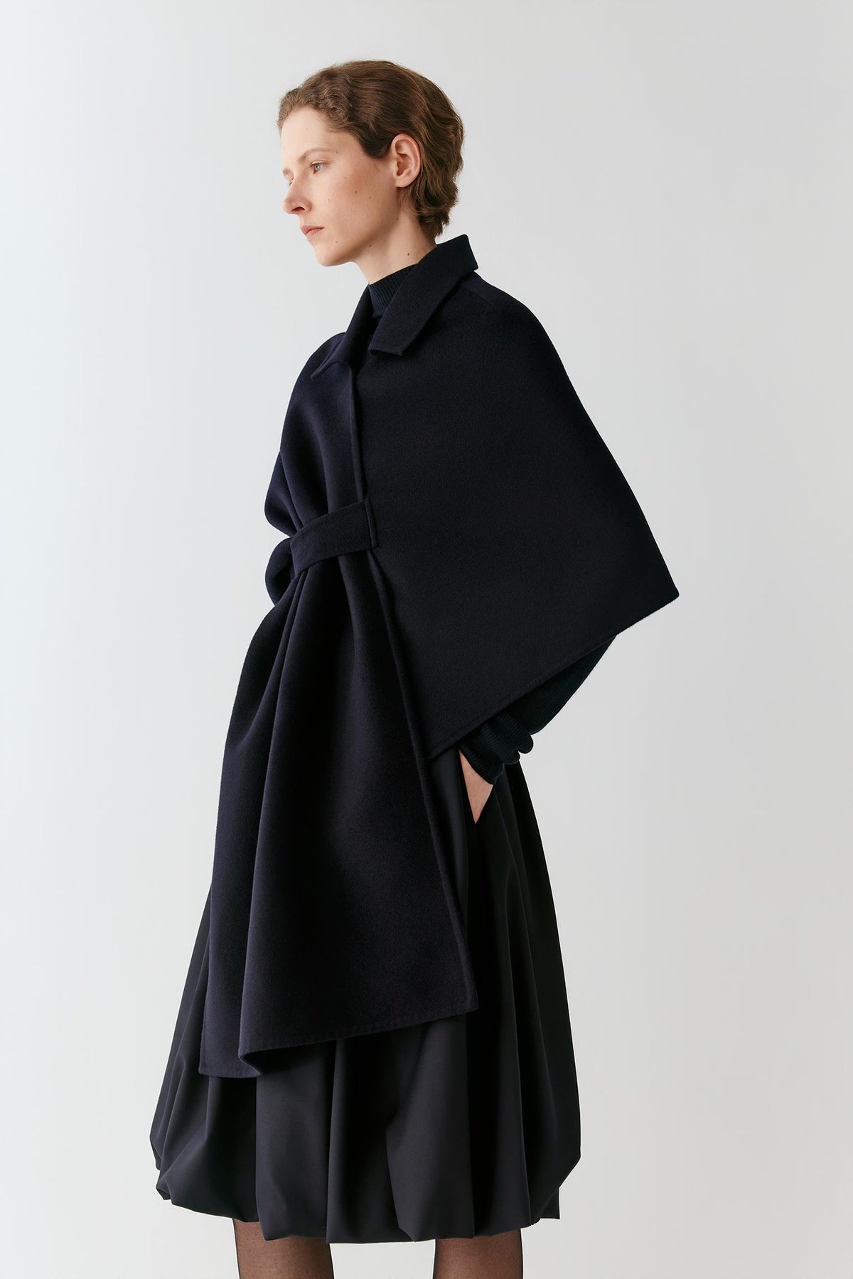 A model wearing a dark navy blue wrap cape with a matching belt and pleated skirt, shown from a side profile against a light gray studio background.