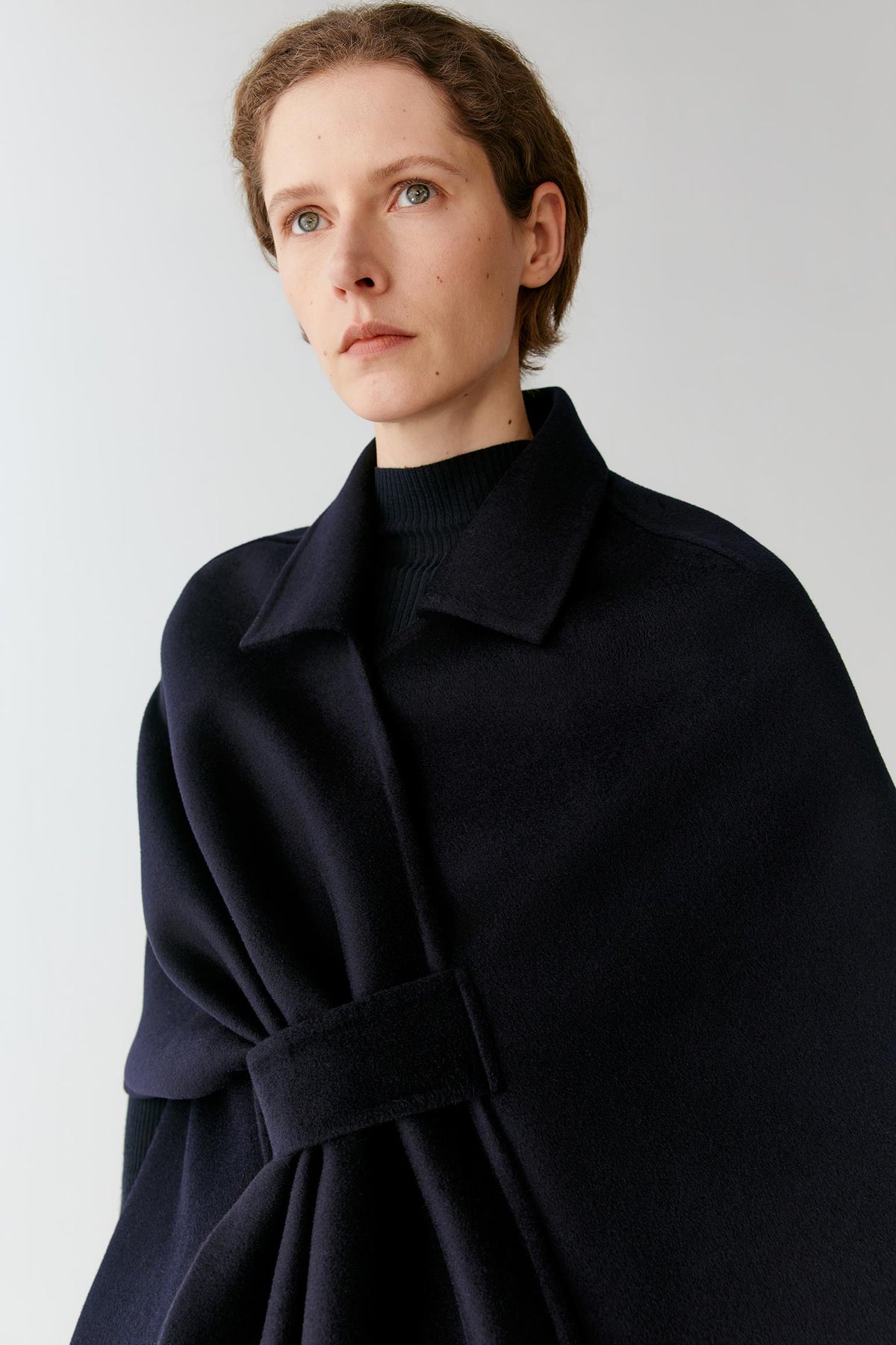 A model wearing a dark navy blue wrap cape with a matching belt over a black ribbed turtleneck, photographed against a neutral light gray background.