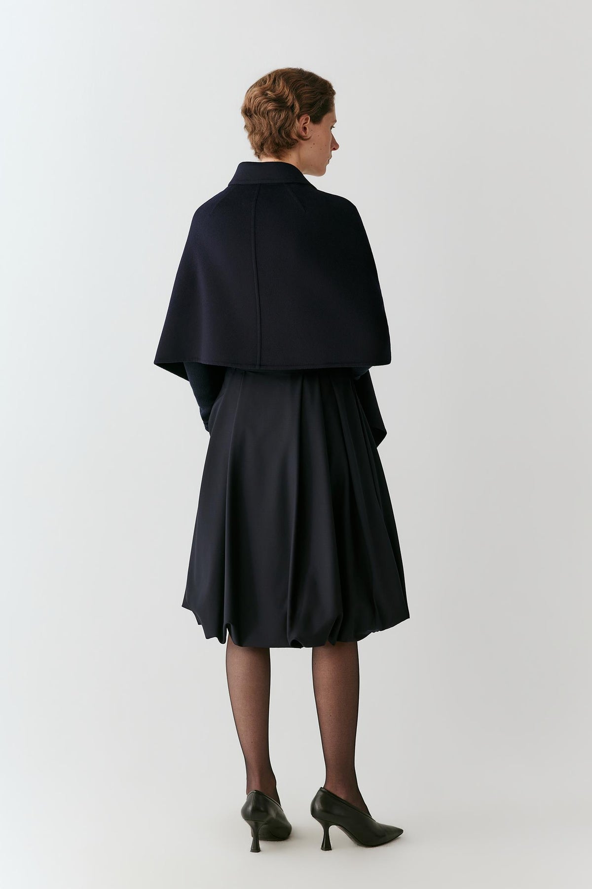 A model shown from the back wearing a dark navy blue cape-style jacket over a matching pleated midi skirt, paired with black tights and black pointed-toe heels, against a light gray studio background.