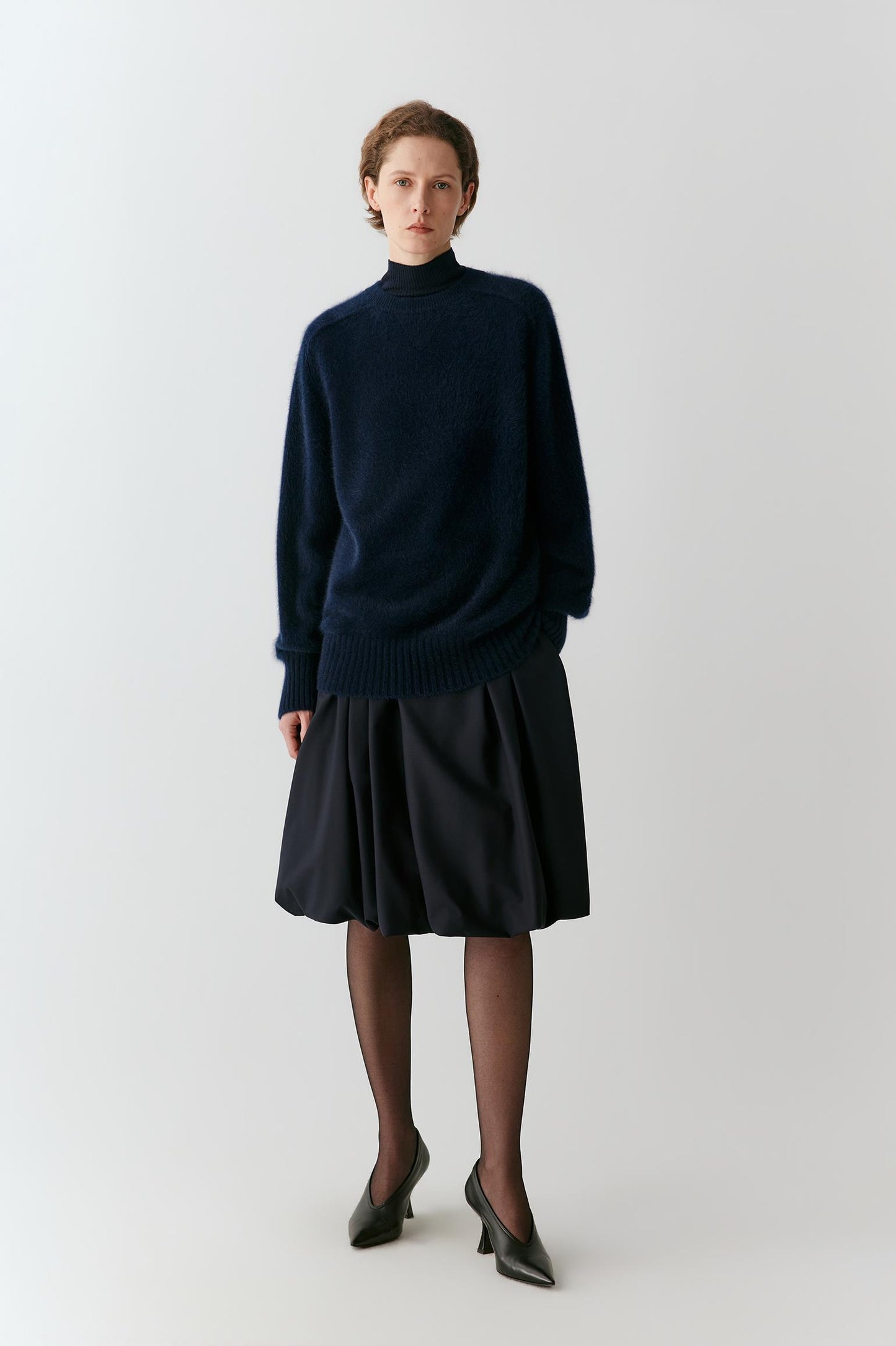 A model stands against a light gray studio background wearing a dark navy blue knit mock-neck sweater and a matching dark navy pleated skirt, paired with sheer black tights and black pointed-toe heels.