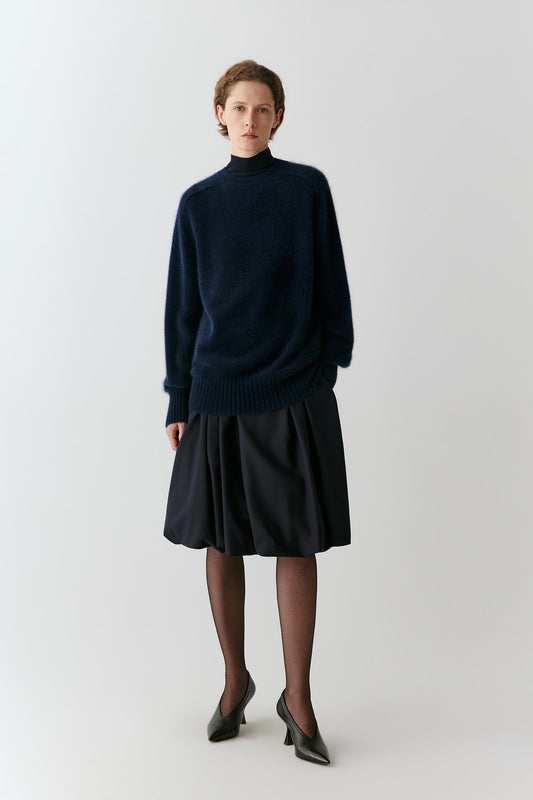 A model stands against a light gray studio background wearing a dark navy blue knit mock-neck sweater and a matching dark navy pleated skirt, paired with sheer black tights and black pointed-toe heels.