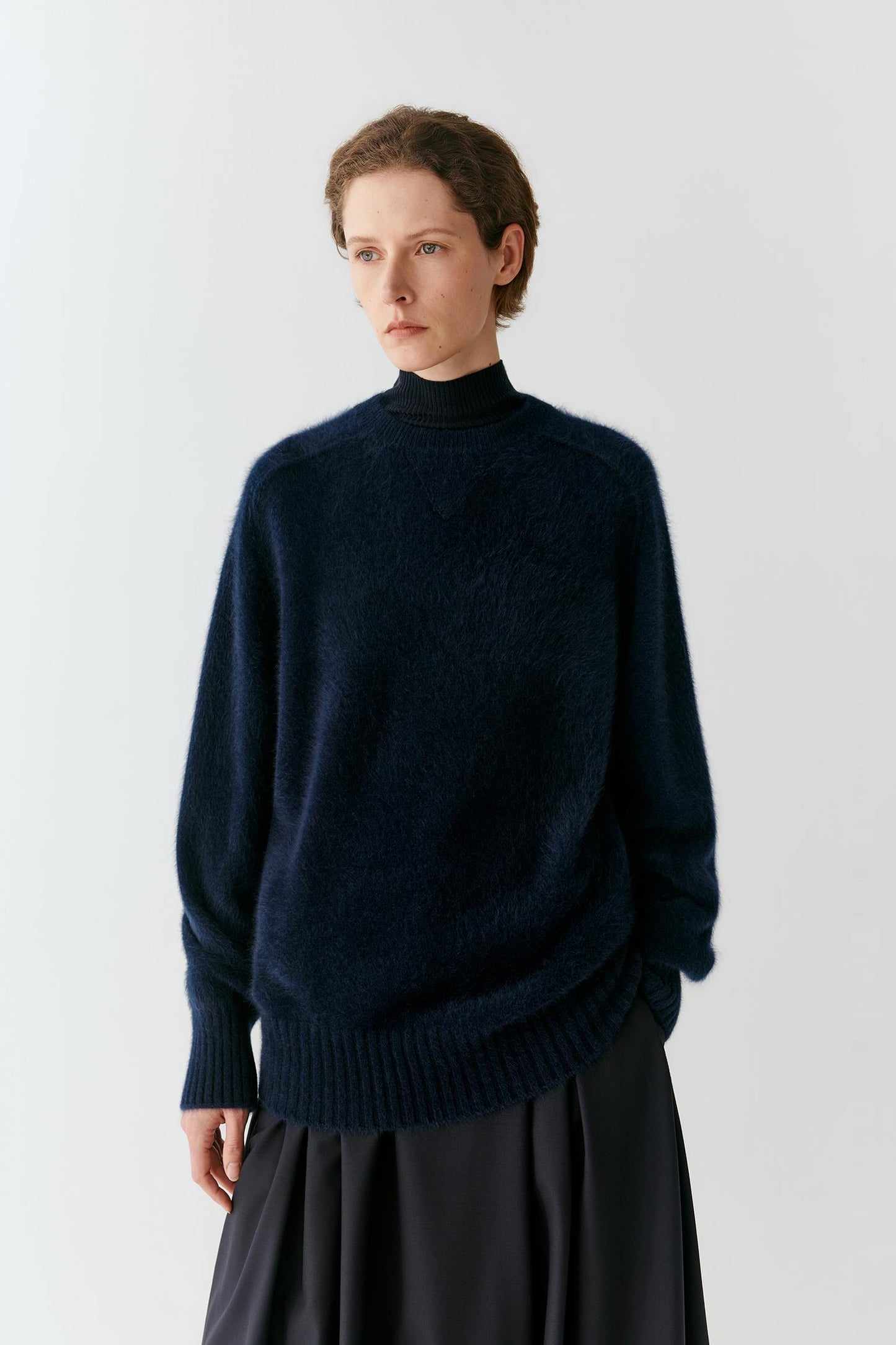 A model wearing a dark navy blue fuzzy knit mock-neck sweater over a black turtleneck, paired with a dark skirt, against a plain light gray background.