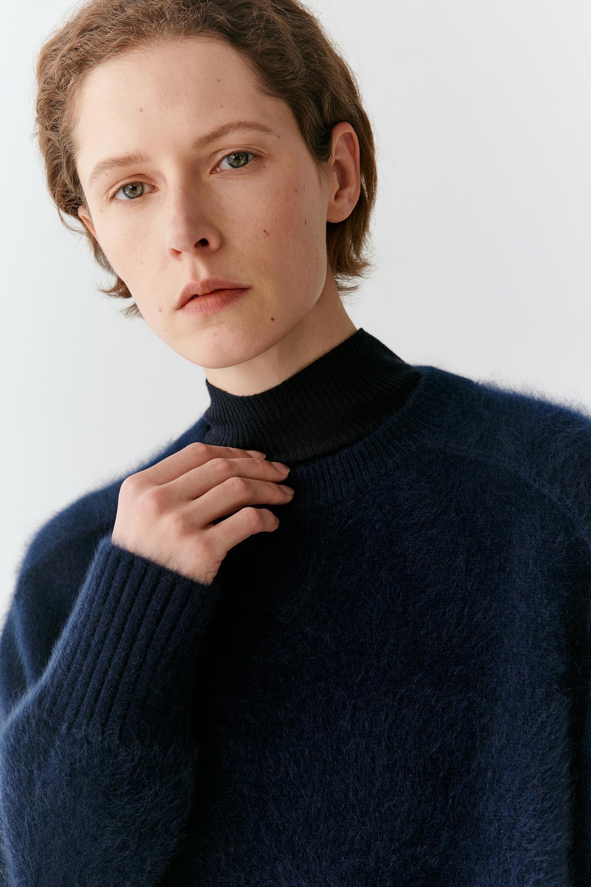 A close-up of a model wearing a dark navy blue fuzzy turtleneck sweater against a plain light gray background.