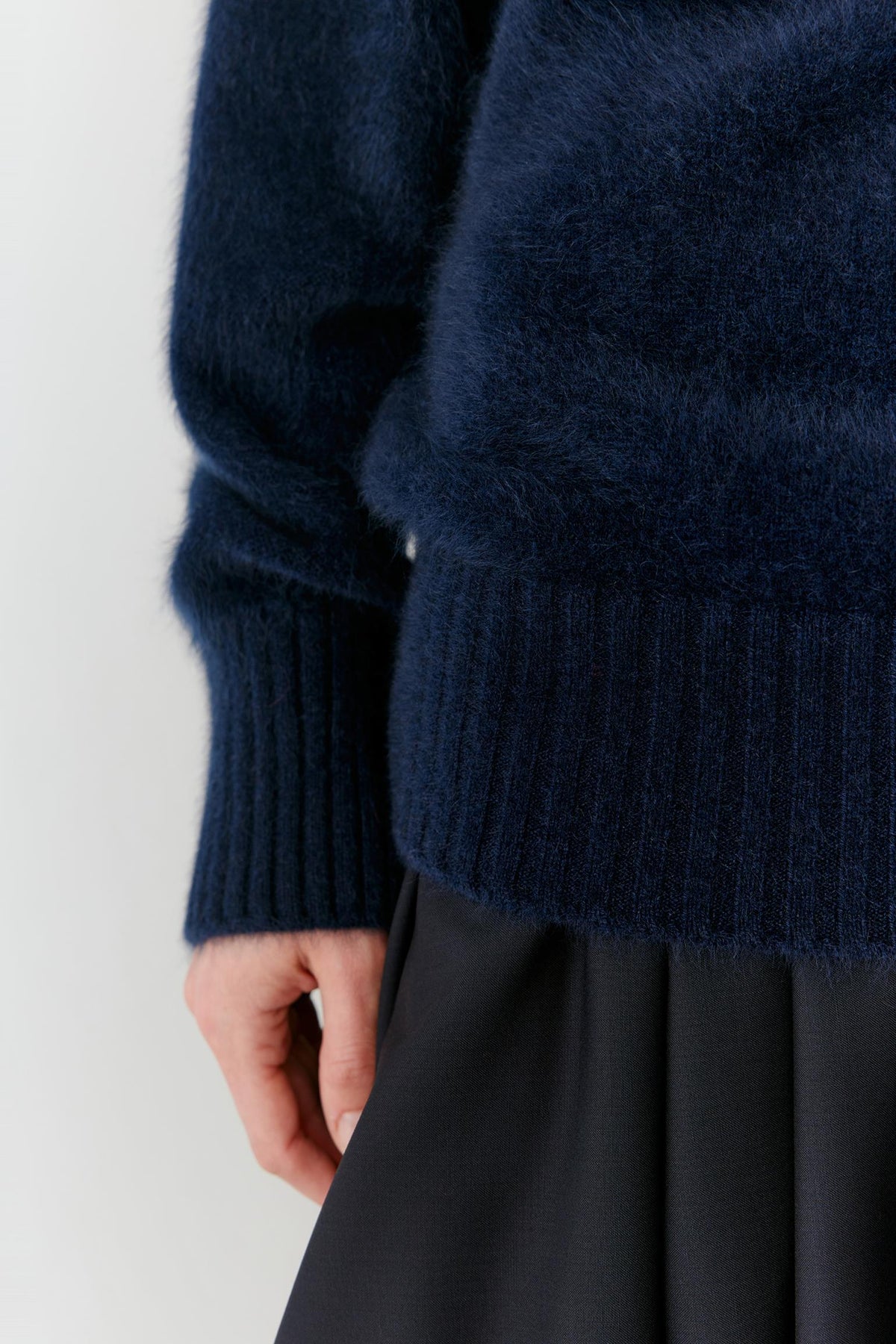 Close-up of a person wearing a dark navy fuzzy knit sweater with a ribbed hem, paired with a dark skirt, against a light gray background.