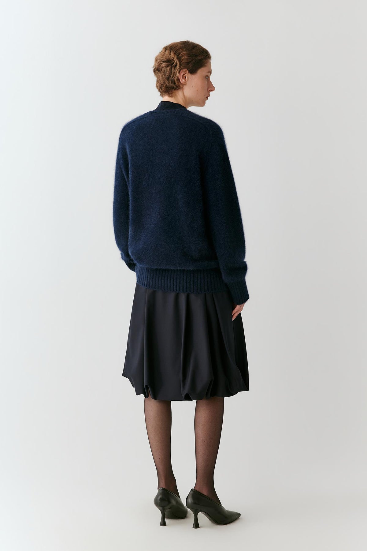 A woman shown from the back wearing a dark navy blue knit sweater and a dark pleated skirt, paired with black tights and black high heels, against a plain light gray studio background.