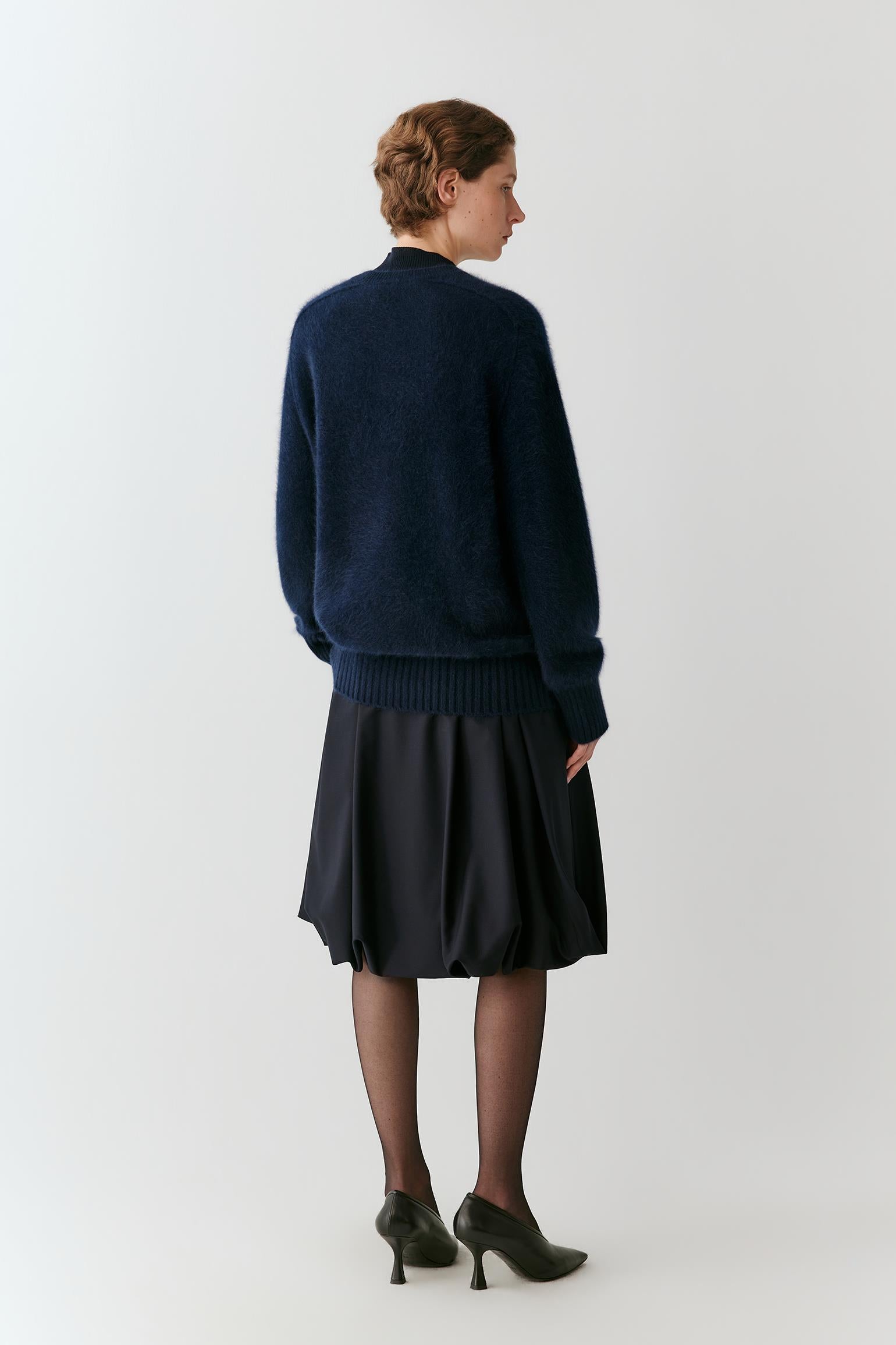 A woman shown from the back wearing a dark navy blue knit sweater and a dark pleated skirt, paired with black tights and black high heels, against a plain light gray studio background.