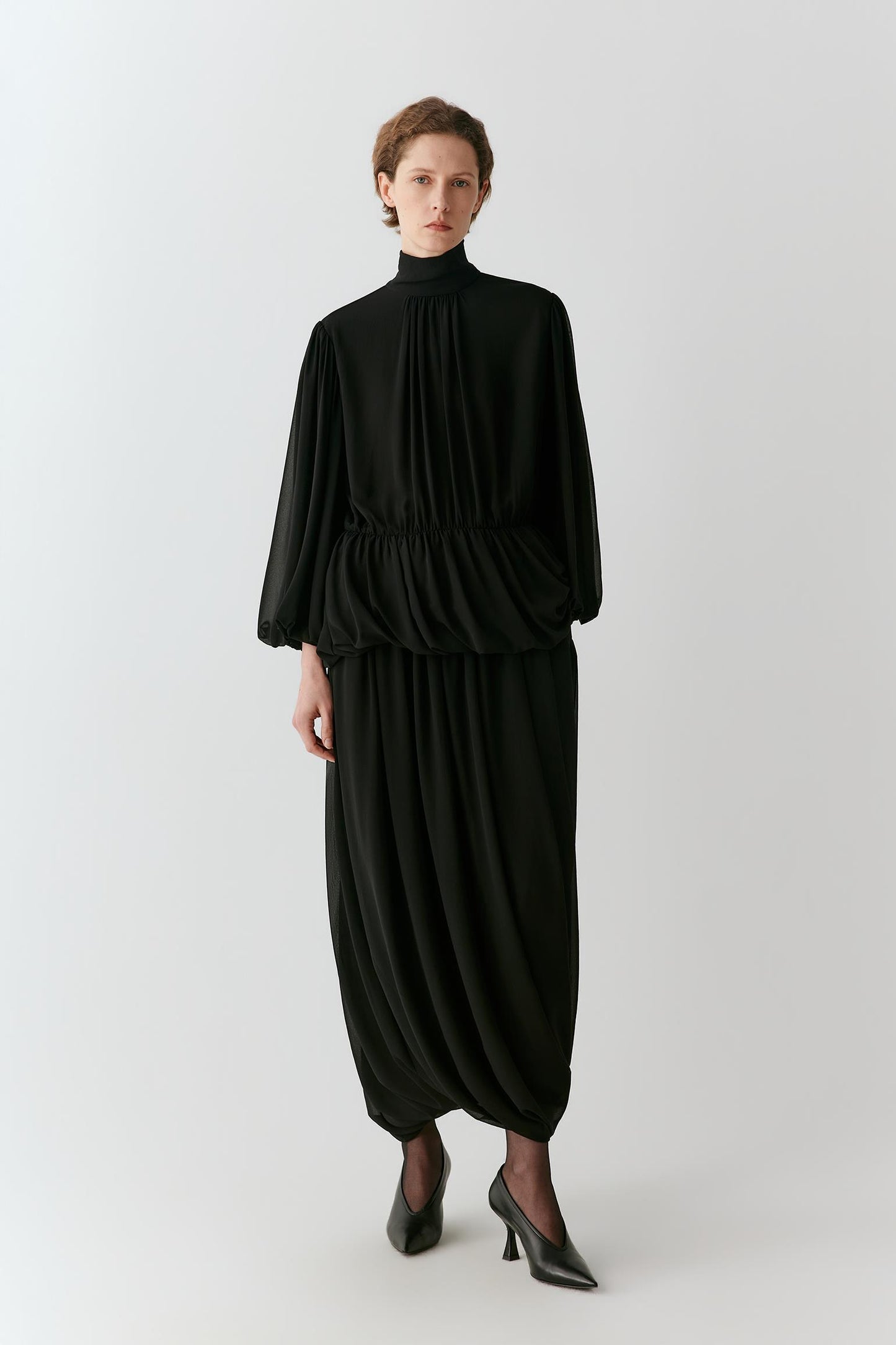 A model wearing a black high-neck, long-sleeved dress with a gathered waist and voluminous draped harem-style legs, paired with black pointed-toe heels, against a light gray studio background.
