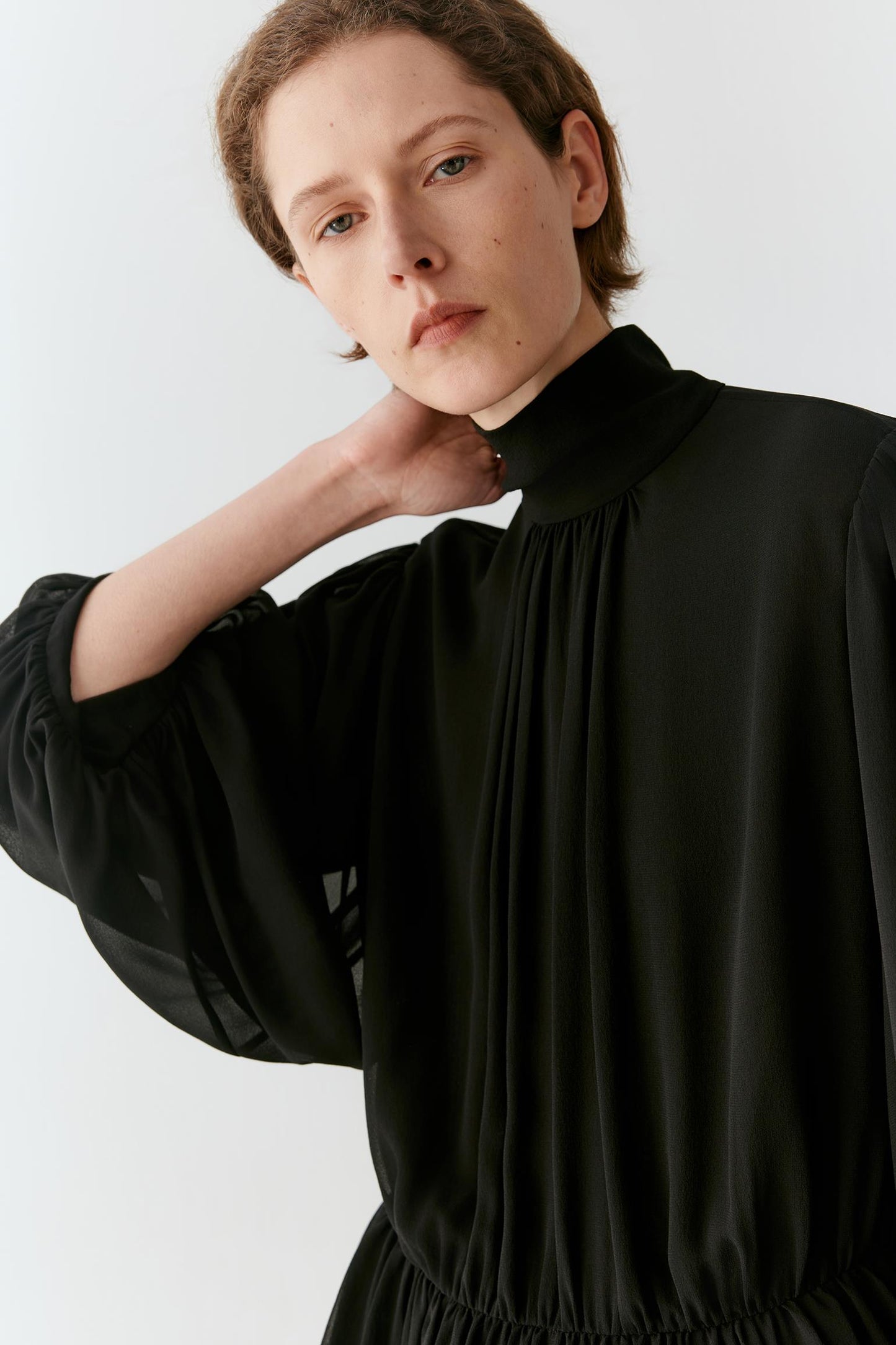 A close-up of a model wearing a black high-neck blouse with voluminous long sleeves and gathered pleats, set against a plain light gray background.