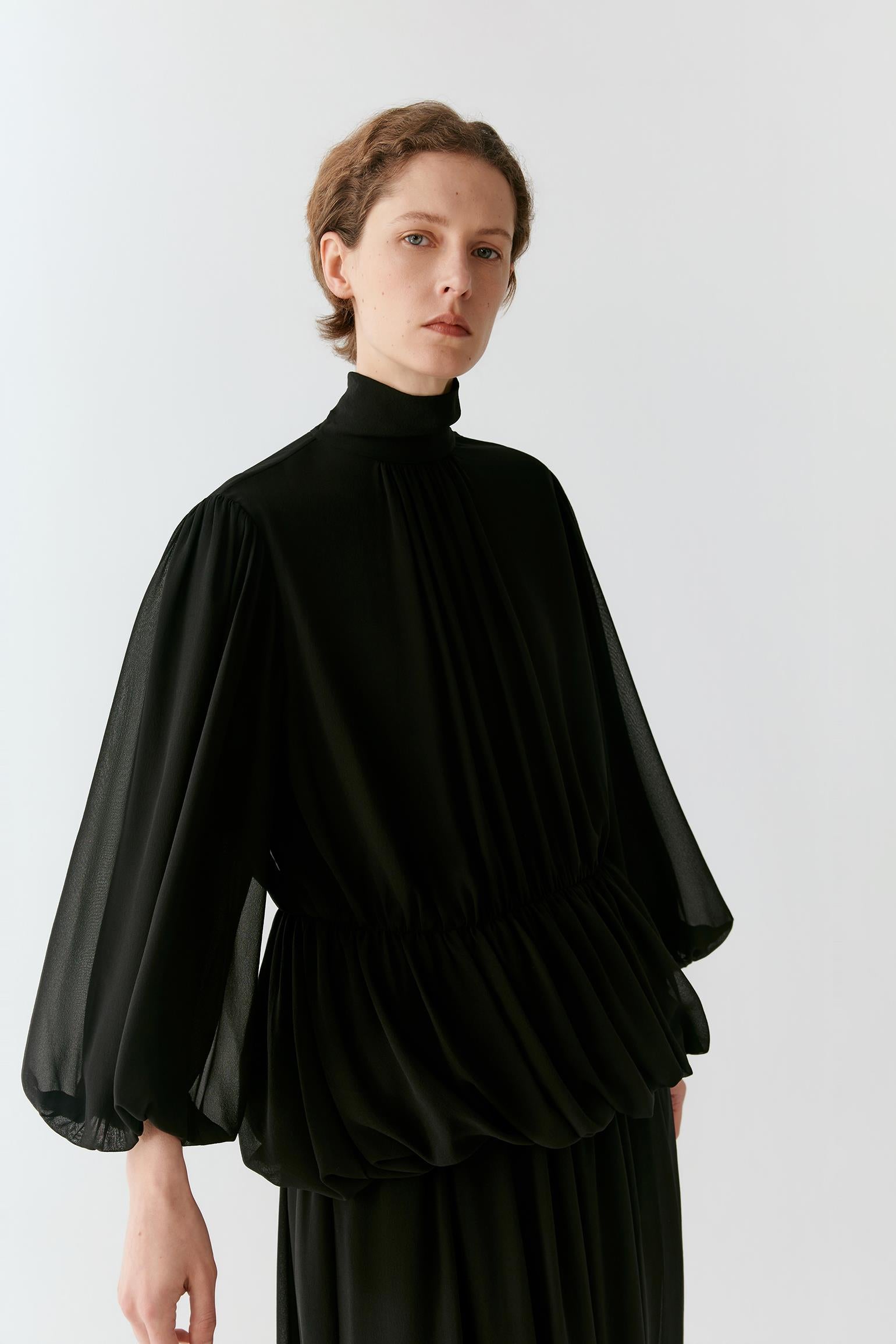 A model wearing a black high-neck blouse with dramatic, voluminous sheer sleeves and a gathered peplum waist, paired with a matching black skirt, set against a plain light gray studio background.