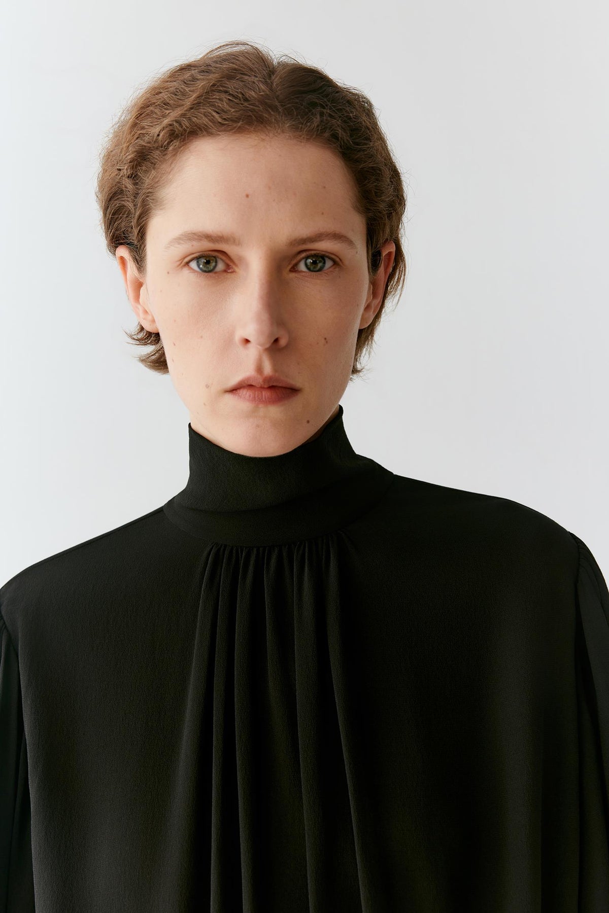 A close-up portrait of a model wearing a black high-neck, long-sleeved blouse with a gathered pleated front, set against a plain light gray background.
