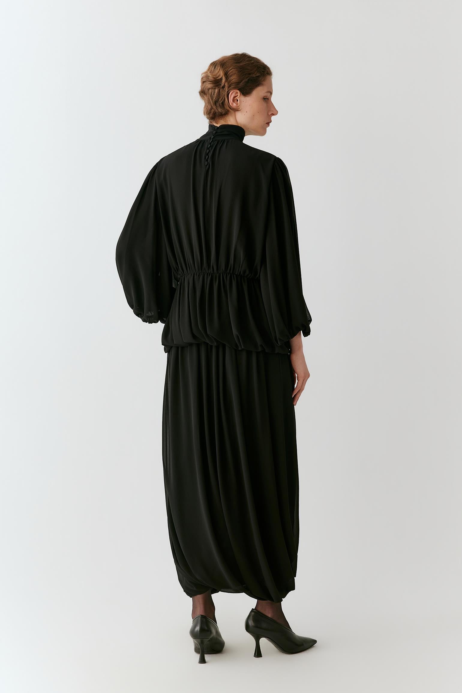 A model is shown from the back wearing a black high-neck, long-sleeved maxi dress with a gathered waist and voluminous balloon sleeves, paired with black pointed-toe heels, against a plain light gray studio background.
