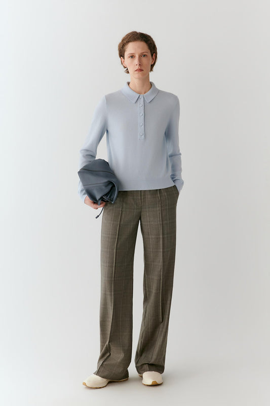 A model stands against a light gray studio background wearing a pale blue long-sleeve polo shirt, olive-brown plaid wide-leg trousers, and cream shoes, while holding a dark blue folded garment.