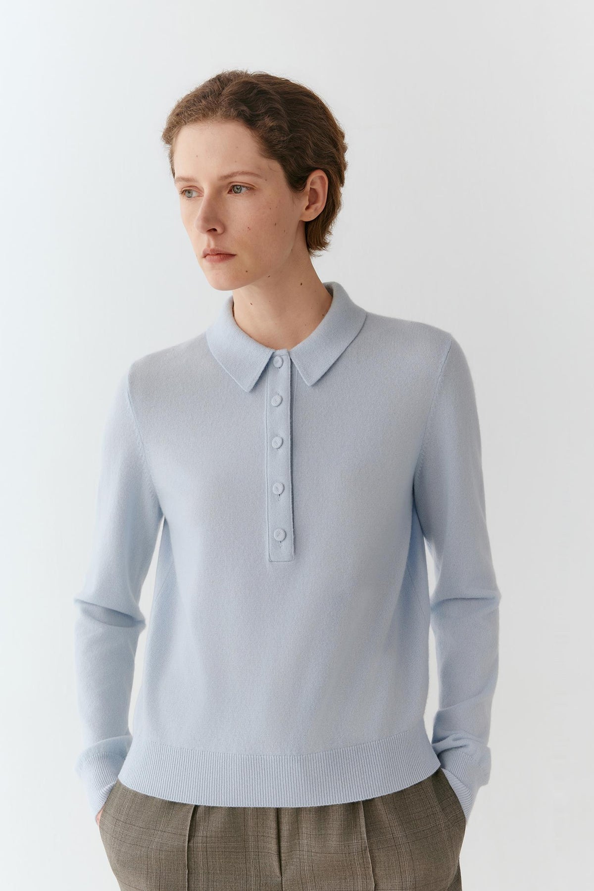 A woman wearing a light blue long-sleeve knit polo sweater with a buttoned collar, paired with brown plaid trousers, photographed against a clean white studio background.