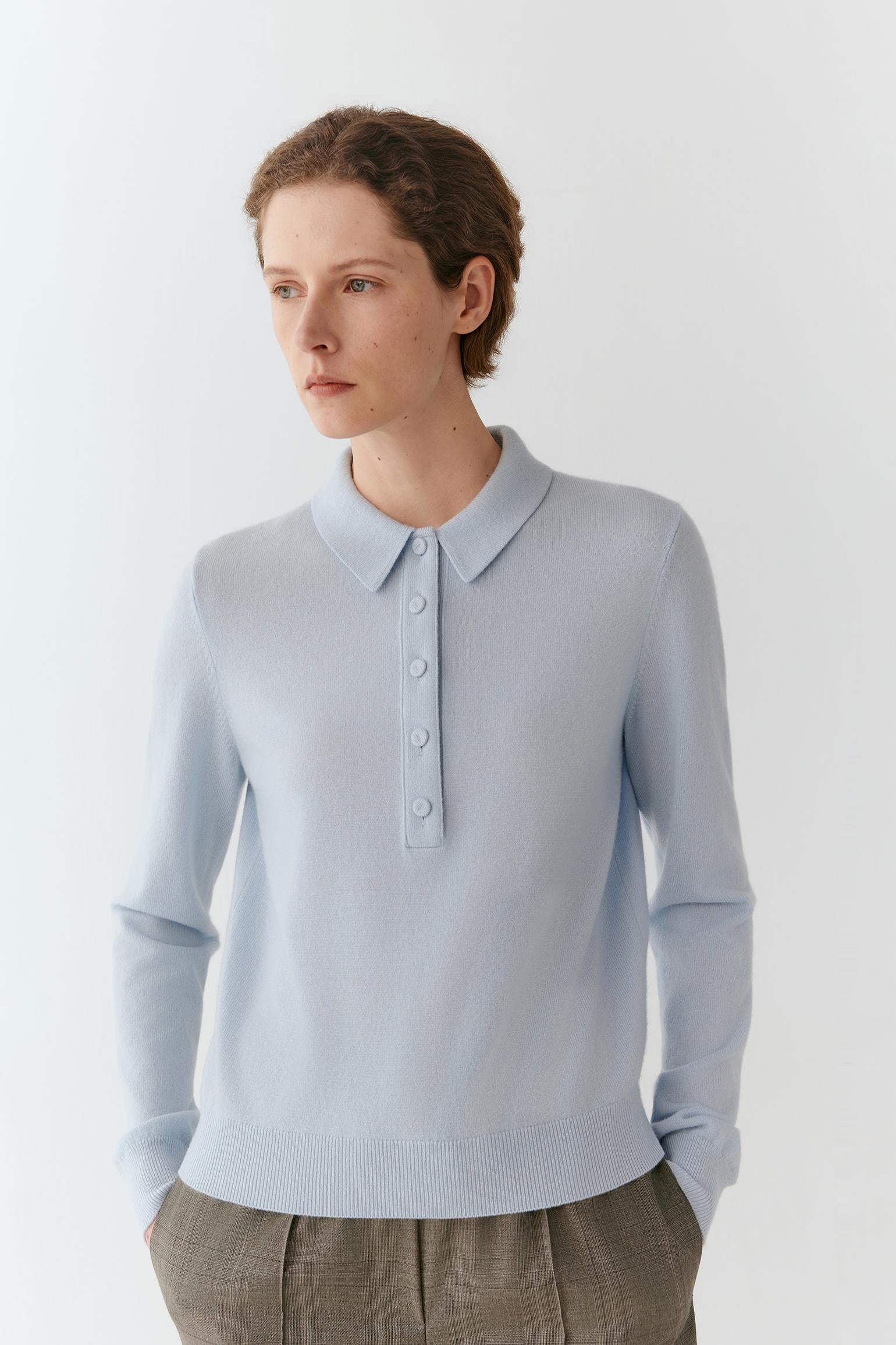 A woman wearing a light blue long-sleeve knit polo sweater with a buttoned collar, paired with brown plaid trousers, photographed against a clean white studio background.