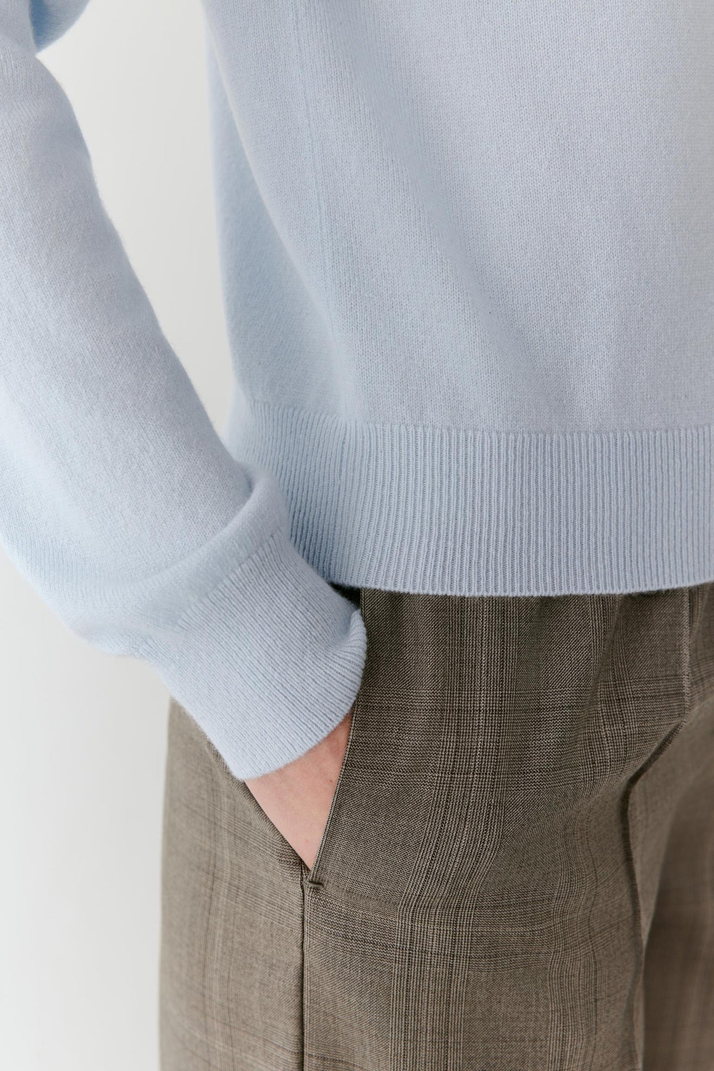 Close-up of a person wearing a light blue ribbed-knit sweater and taupe plaid trousers, with one hand in a pocket.