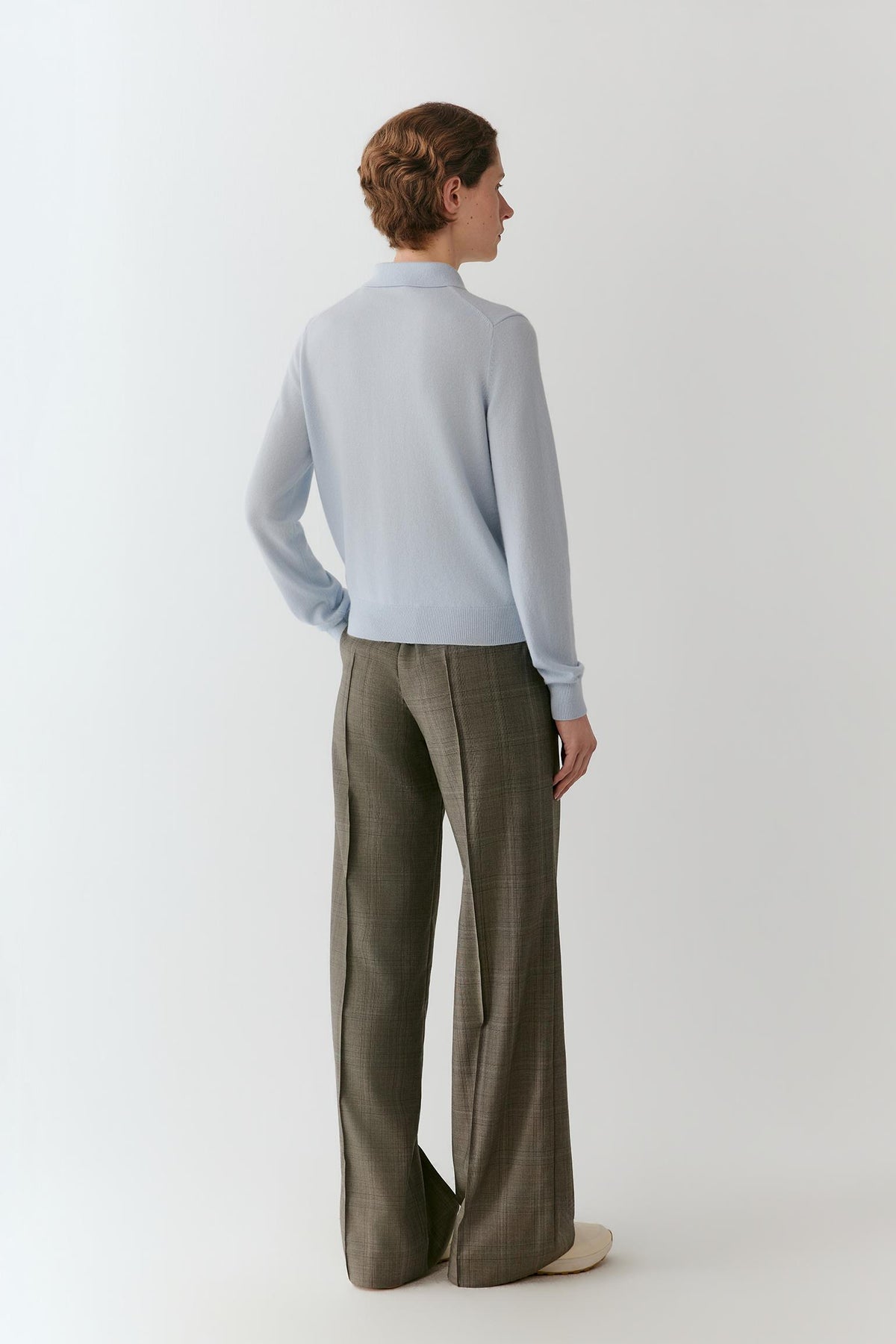 A woman is shown from the back wearing a light blue mock-neck knit sweater and wide-leg brown plaid trousers, standing against a plain light gray studio background.
