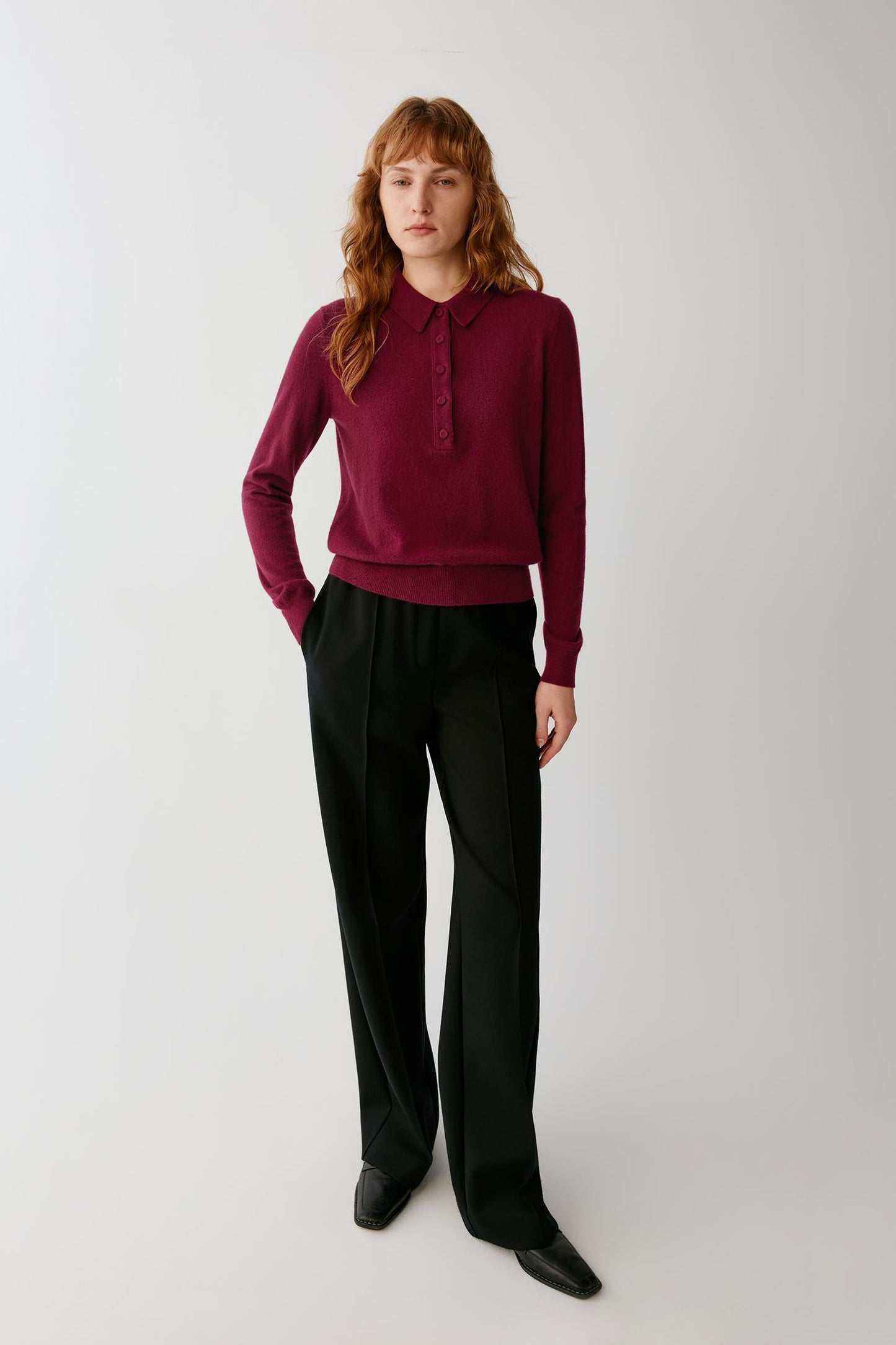 A woman stands against a light gray studio background wearing a burgundy long-sleeve knit polo-style top with black wide-leg trousers and black pointed shoes.