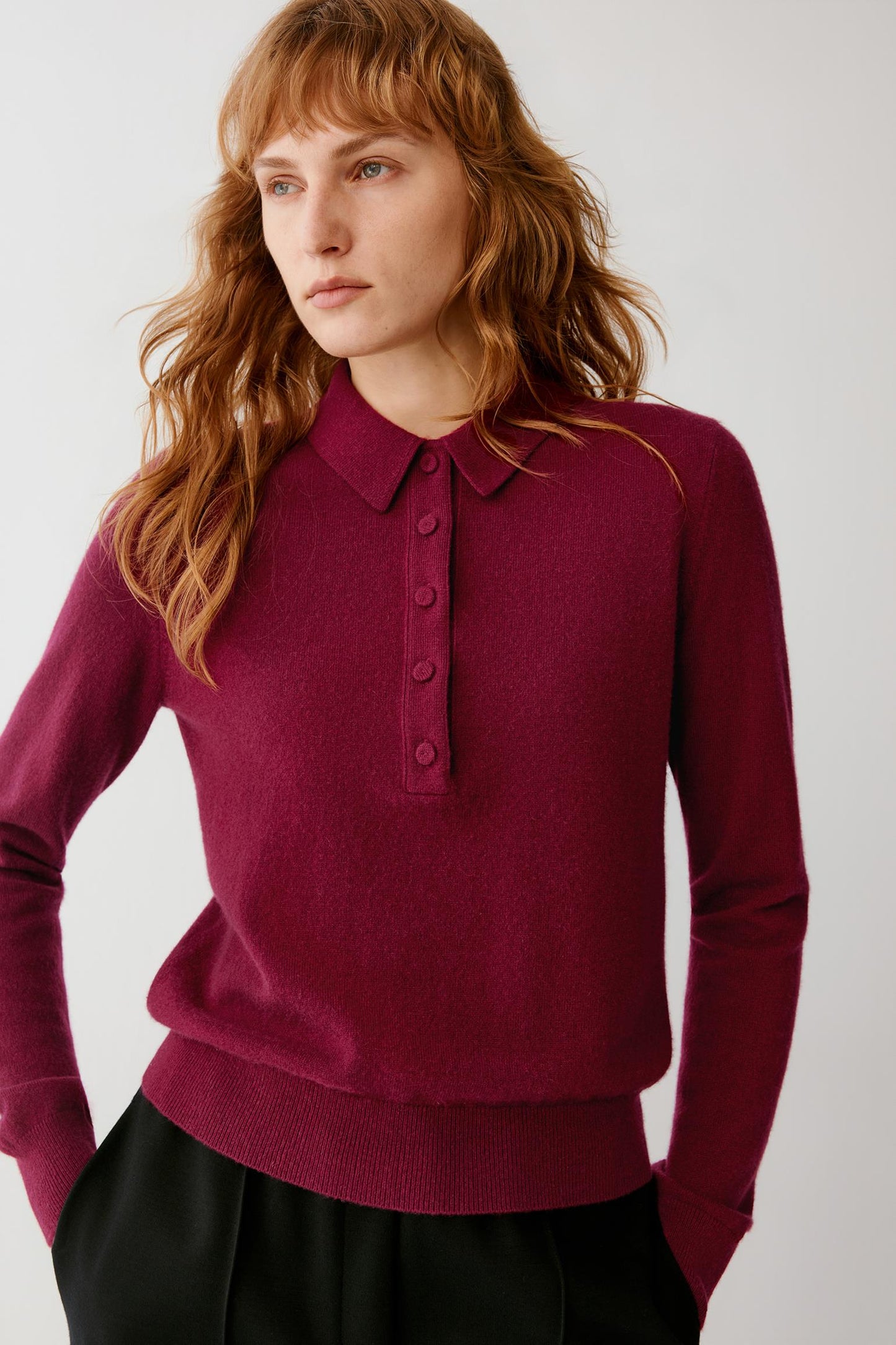 A woman wearing a long-sleeve polo-style knit sweater in deep burgundy with a buttoned collar, paired with black bottoms, against a plain light gray studio background.