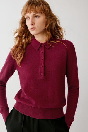A woman wearing a long-sleeve polo-style knit sweater in deep burgundy with a buttoned collar, paired with black bottoms, against a plain light gray studio background.