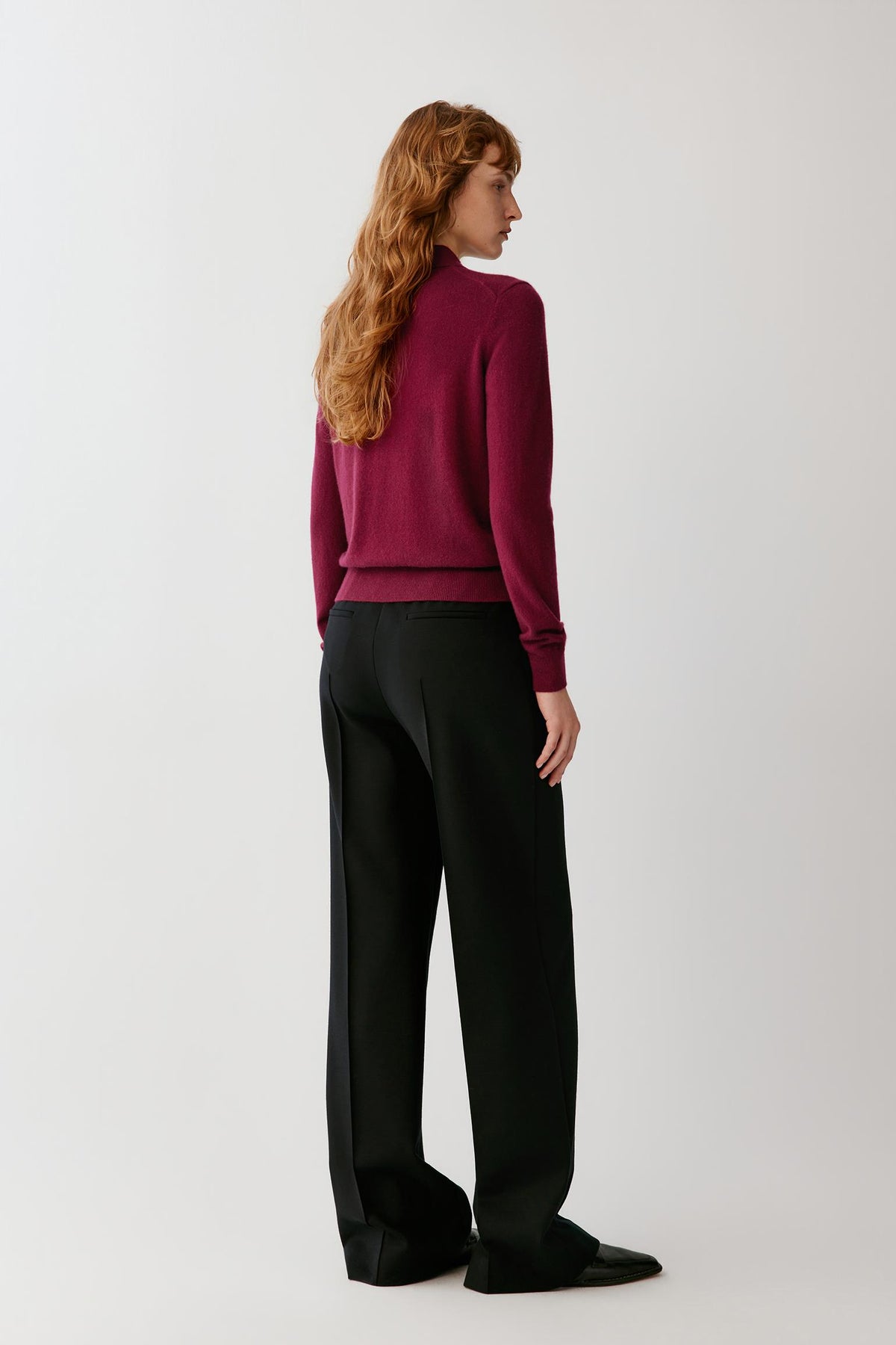 A woman with long wavy red hair is shown from the back wearing a burgundy knit sweater and black wide-leg trousers against a plain light gray studio background.