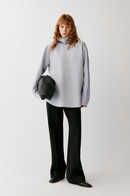 A model stands against a light gray studio background wearing an oversized light gray mock-neck top, black wide-leg trousers, black pointed shoes, and holding a black textured pouch bag.