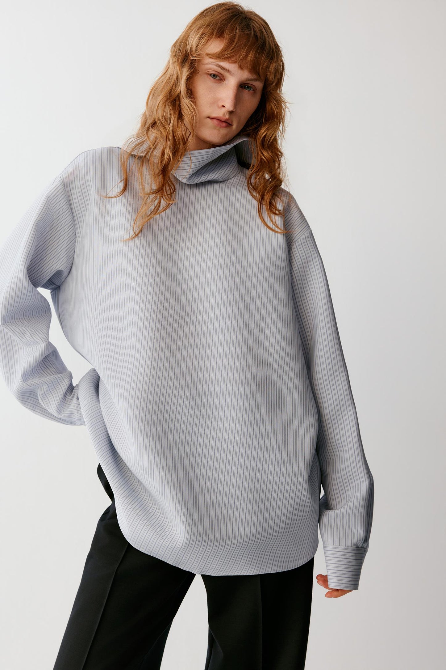 A woman wearing a light gray ribbed knit turtleneck sweater and black pants, photographed against a plain light gray studio background.