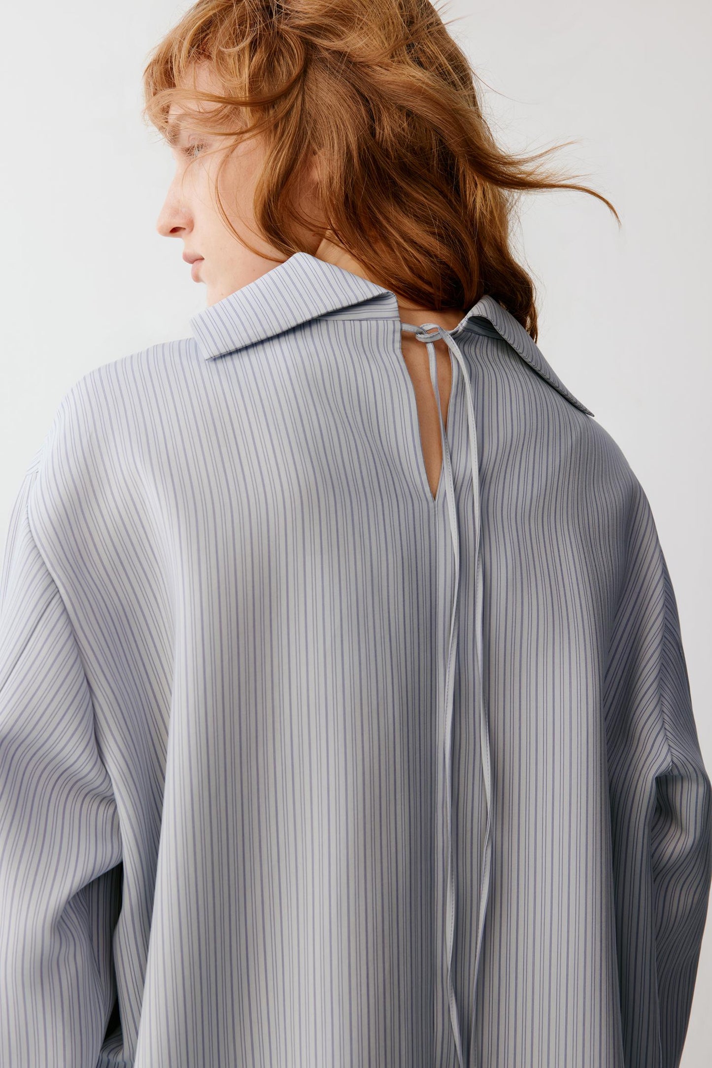 A woman shown from the back wearing a loose, light gray and white vertically striped blouse with a tie detail at the neckline, against a plain light background.