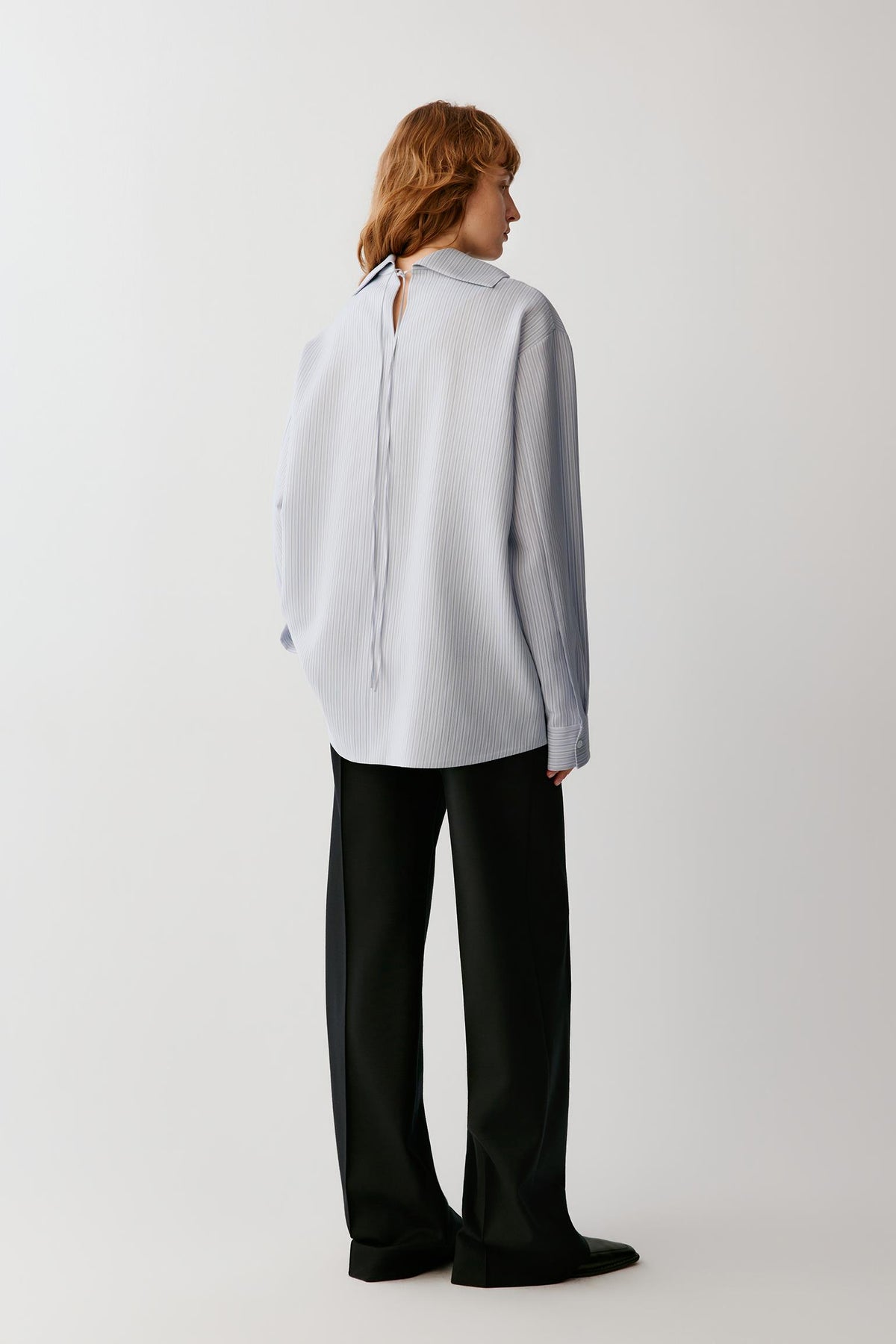 A model shown from the back wearing a light gray long-sleeve blouse with a tie-neck detail and black wide-leg trousers, photographed against a plain light gray studio background.