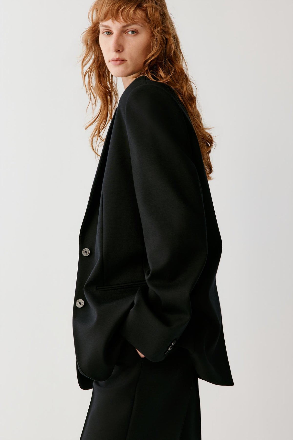A model with long wavy red hair is shown from a side-back angle wearing a black oversized blazer with silver buttons, set against a plain light gray studio background.