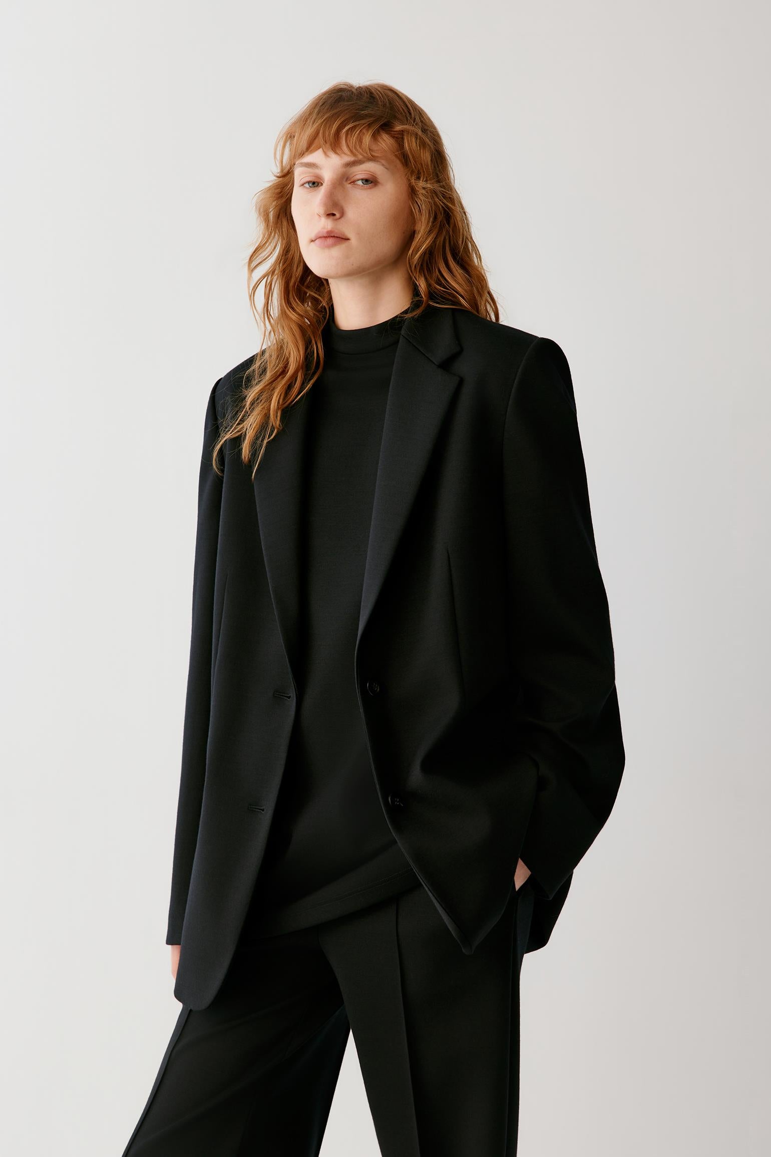 A model with wavy red hair wearing an oversized black blazer over a black mock neck top and matching black trousers, photographed against a light gray studio background.