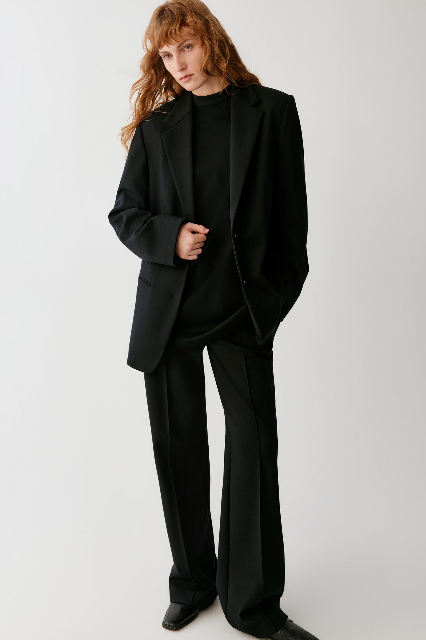 A model with red hair wearing a black oversized blazer, black high-neck top, black wide-leg trousers, and black shoes, standing against a light gray studio background.