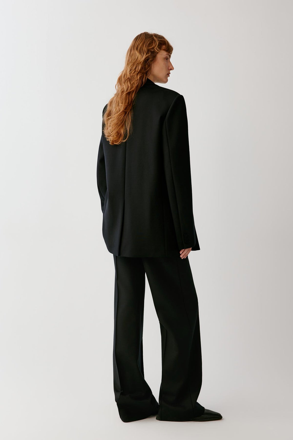 A model with long wavy red hair is shown from the back wearing a black wide-leg blazer and matching wide-leg trousers against a light gray studio background.