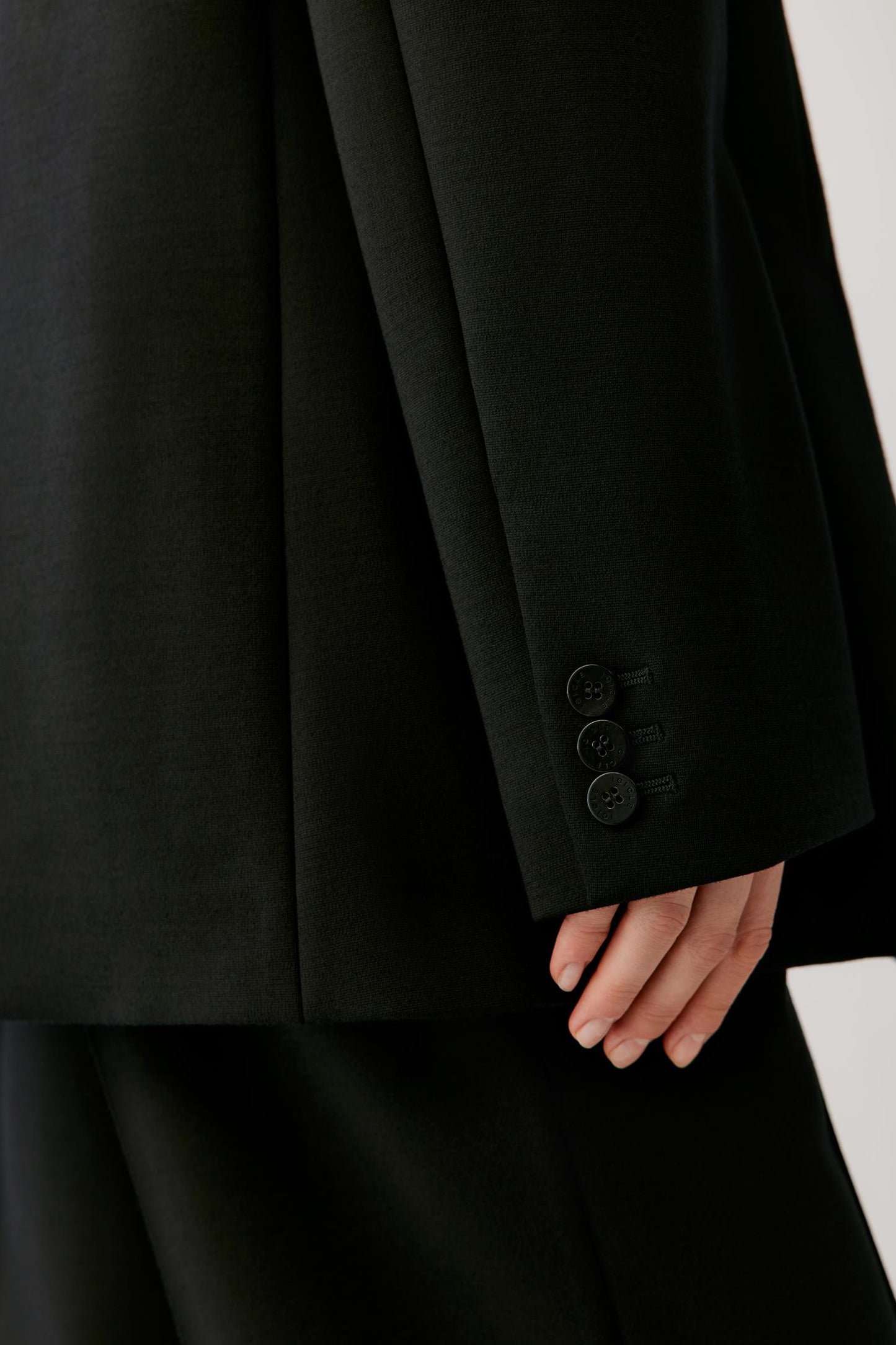 Close-up of a black tailored jacket sleeve with three buttons and a hand resting near the cuff, against a light neutral background.