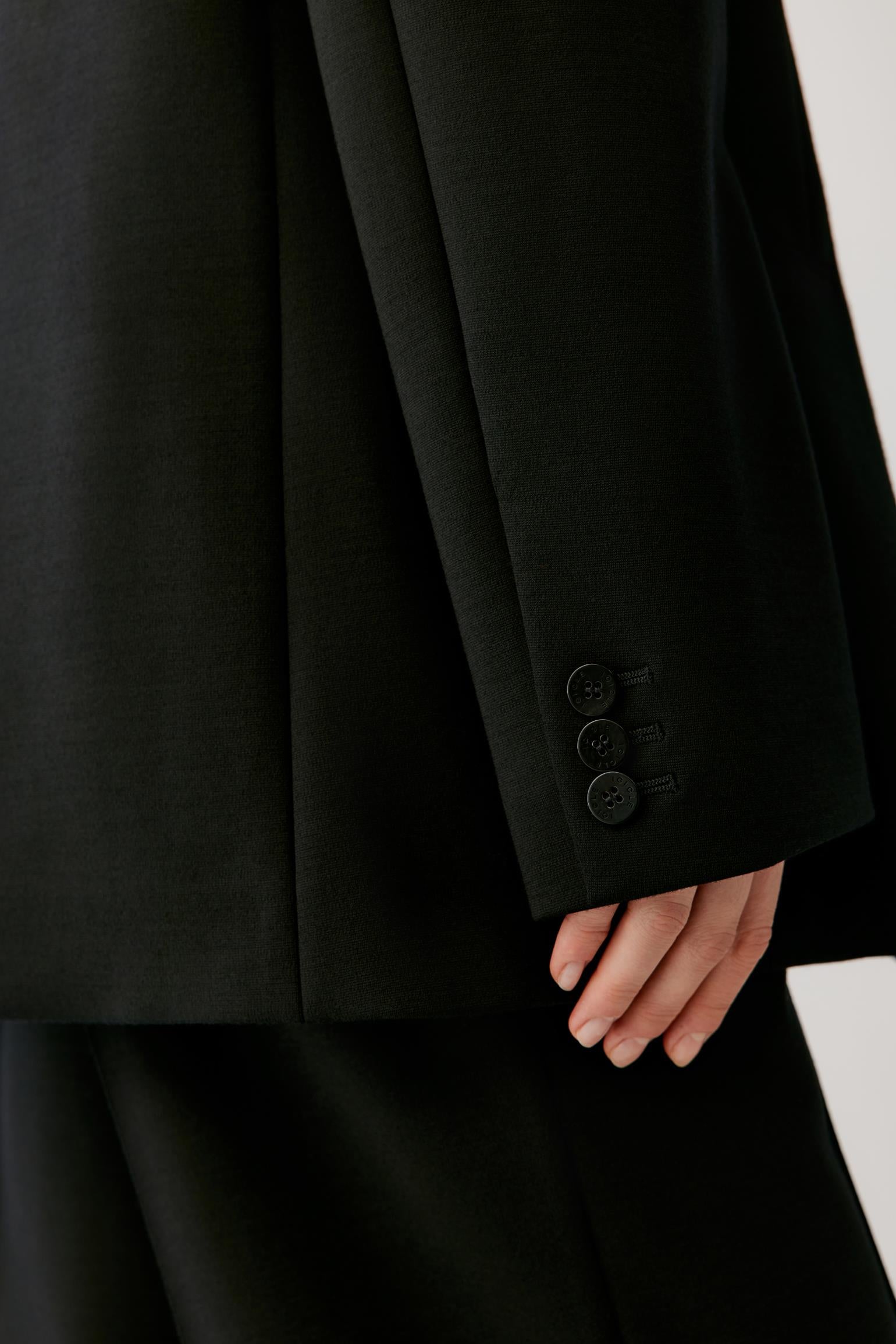 Close-up of a black tailored jacket sleeve with three buttons and a hand resting near the cuff, against a light neutral background.