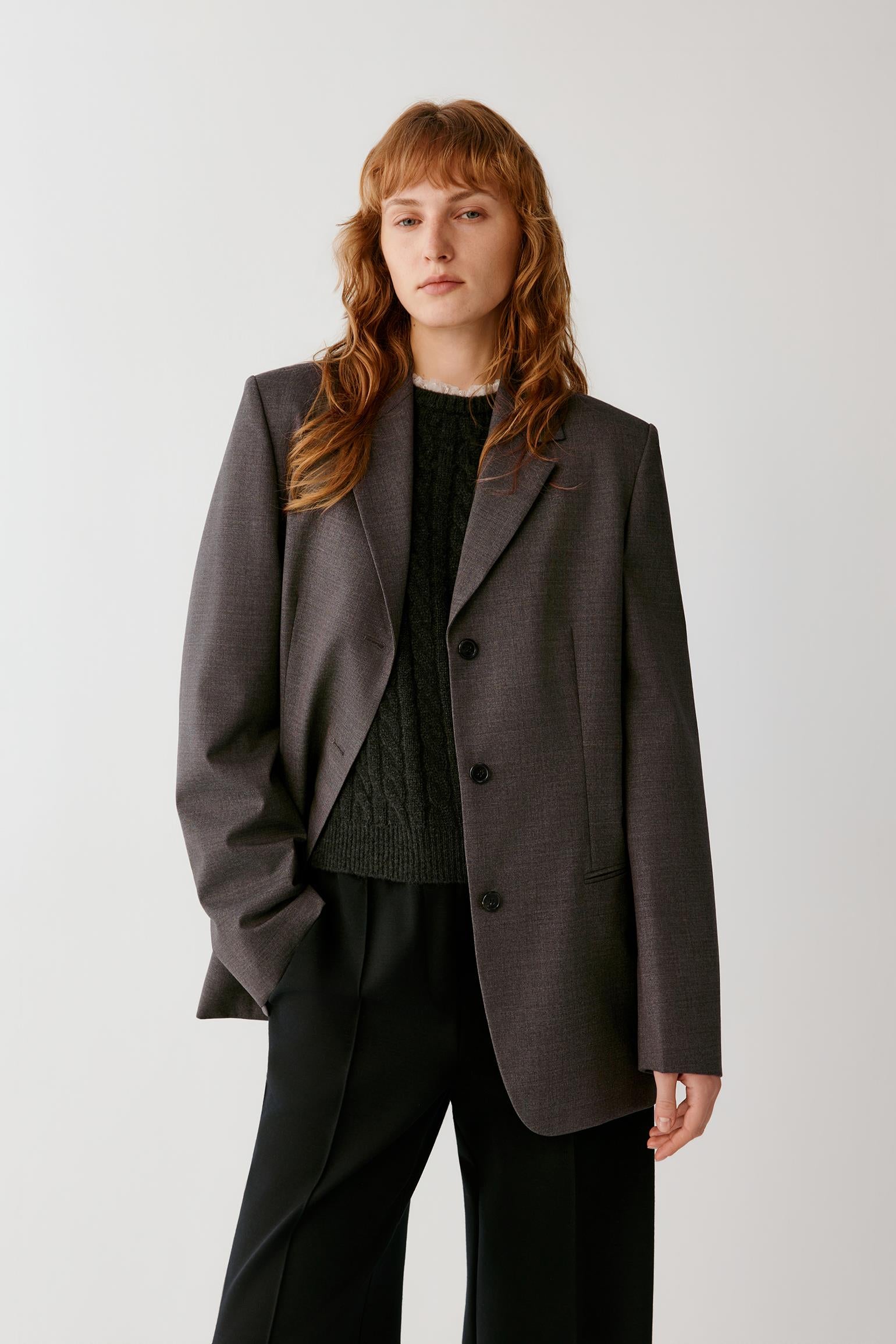 Front view of a woman wearing a grey melange wool blazer, highlighting the straight silhouette, notched collar and three-button fastening.
