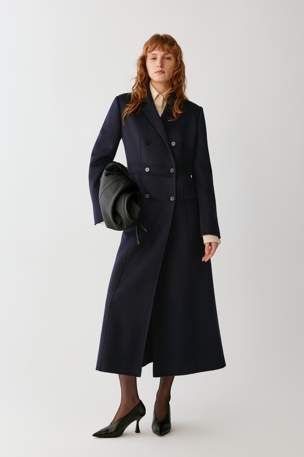 A woman stands against a light gray studio background wearing a long navy double-breasted coat over a cream collared blouse, holding a black clutch, with black pointed heels.