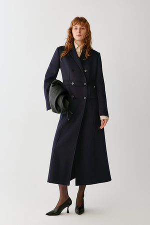 A woman stands against a light gray studio background wearing a long navy double-breasted coat over a cream collared blouse, holding a black clutch, with black pointed heels.