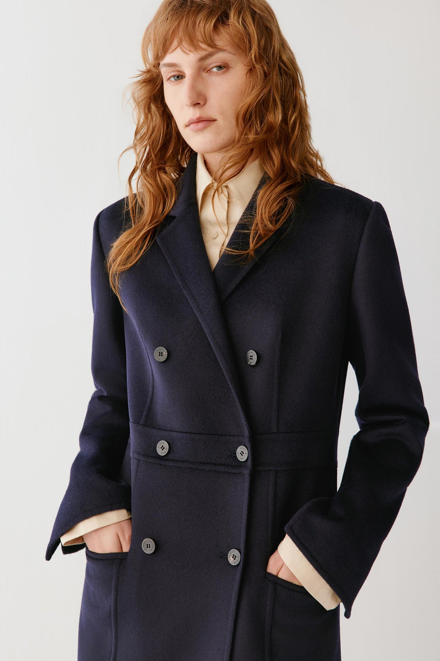 A woman wearing a dark navy double-breasted coat over a cream shirt, photographed against a plain light gray studio background.