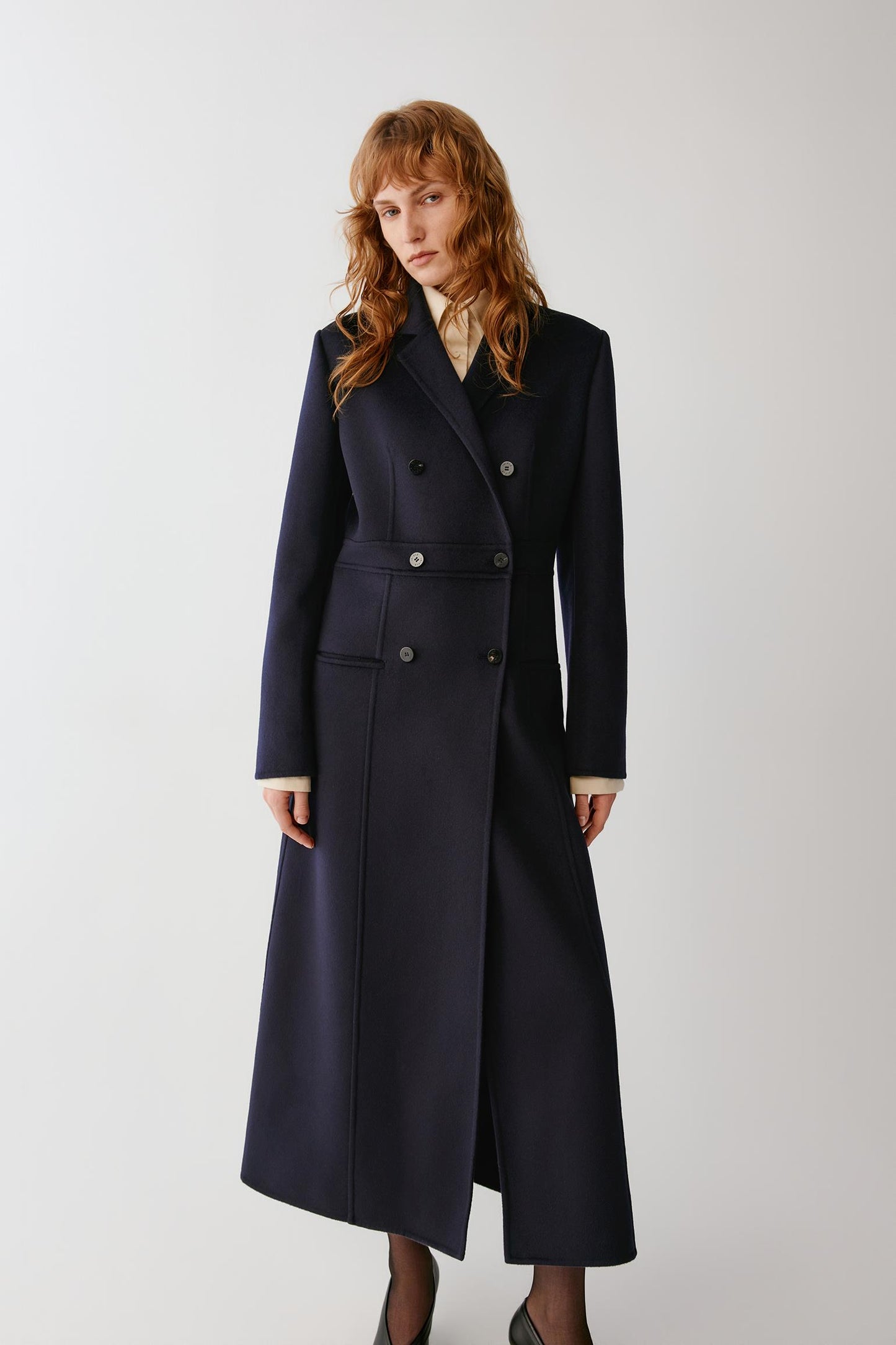 A woman modeling a long navy double-breasted coat with a belted waist, shown against a plain light gray studio background.