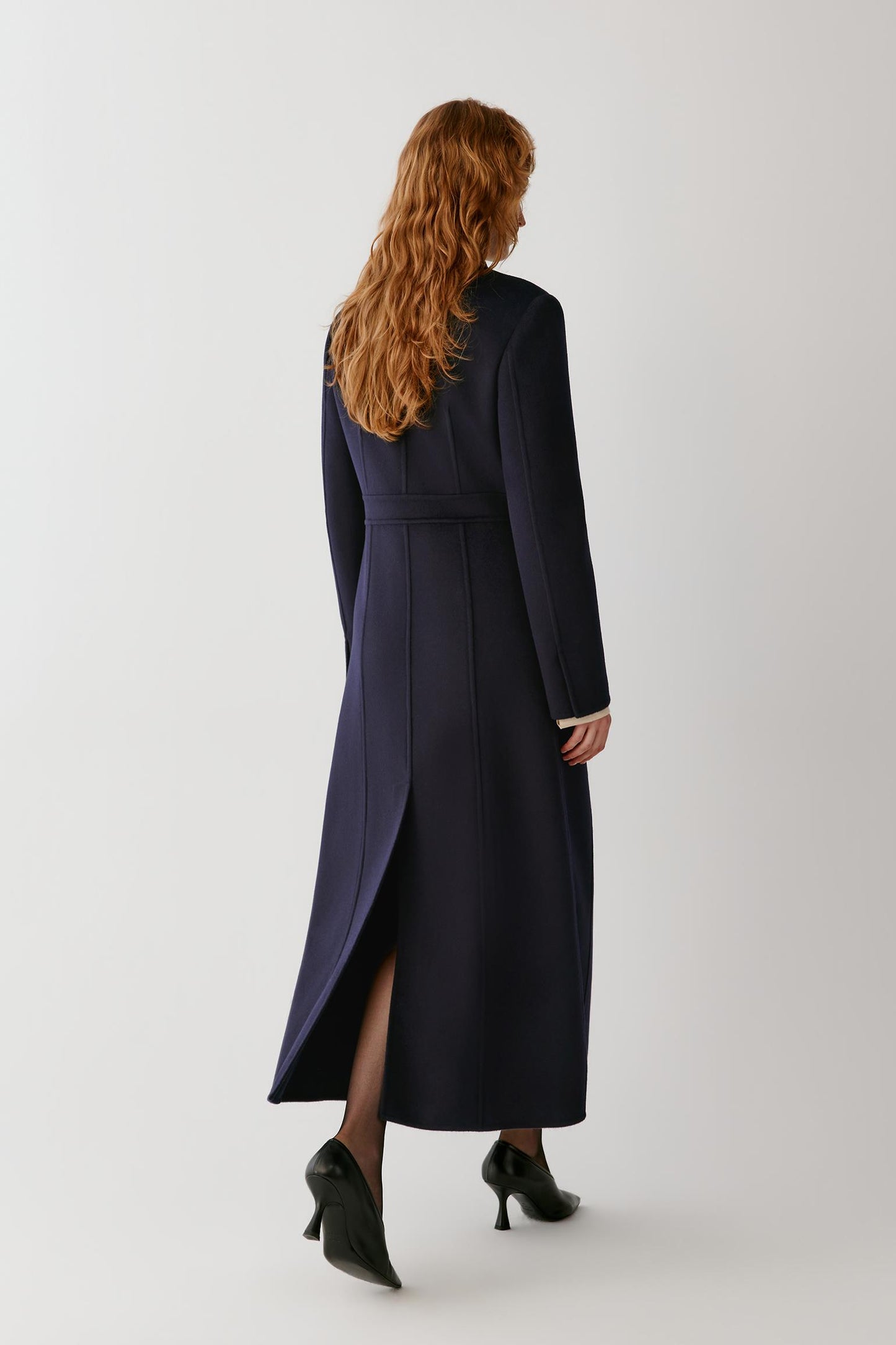 A woman with long wavy red hair is shown from behind wearing a long navy blue wrap coat with a waist belt, black tights, and black pointed-toe heels against a light gray studio background.