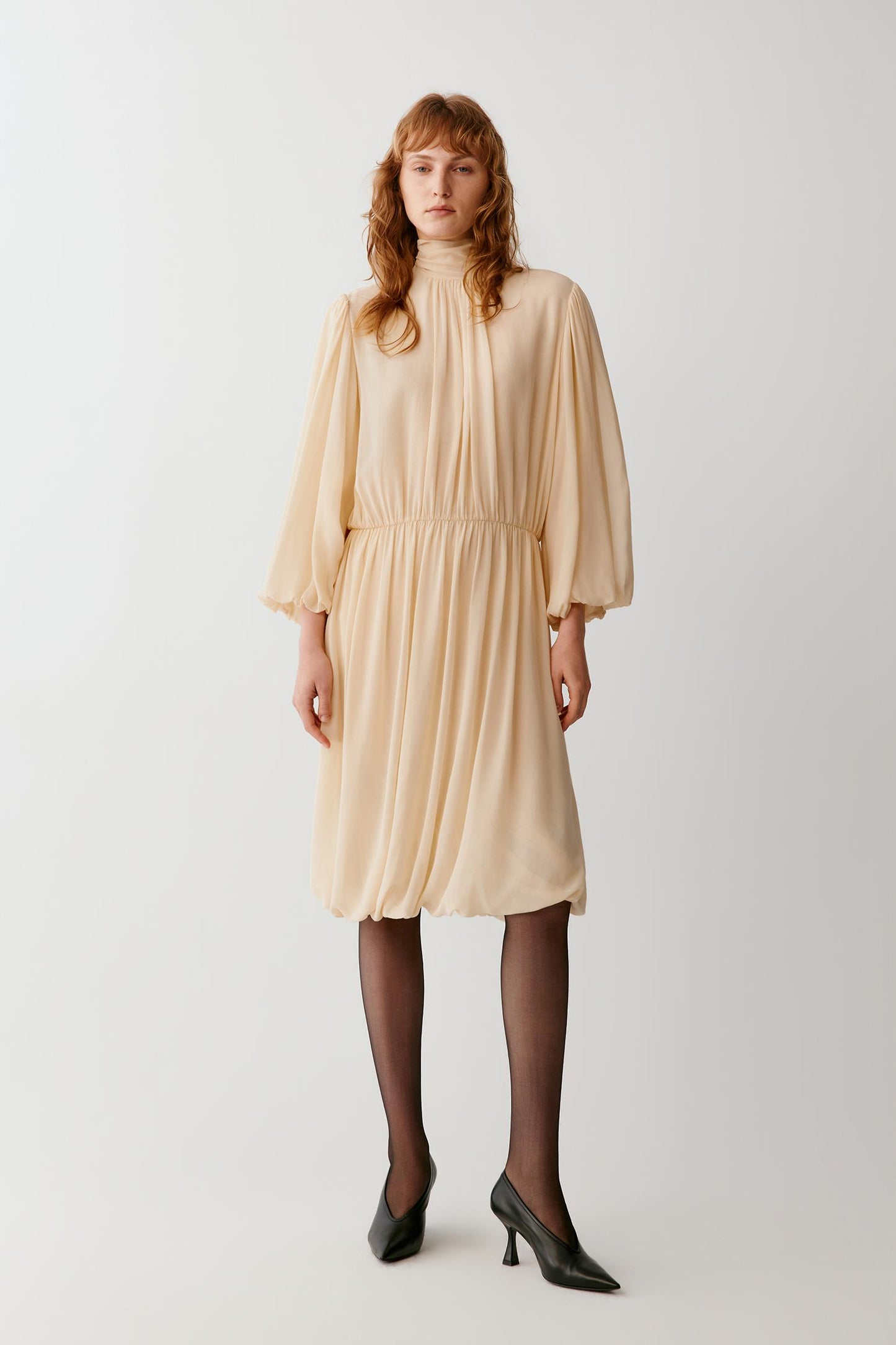 A woman modeling a light beige high-neck midi dress with voluminous three-quarter sleeves and a gathered waist, paired with black tights and black pointed-toe heels, against a plain light gray studio background.