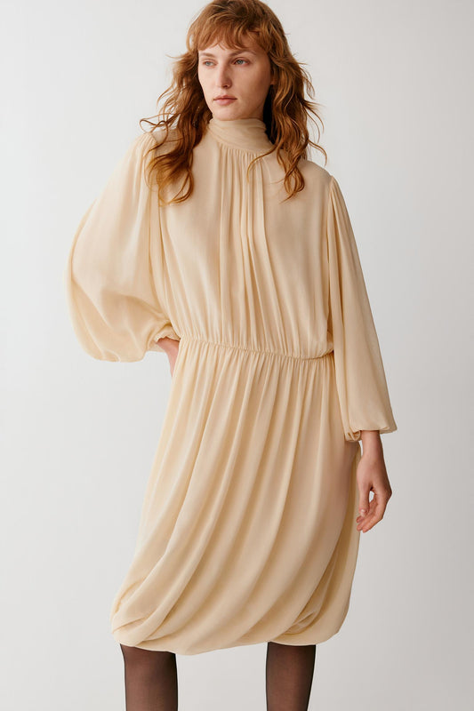 A woman modeling a light beige high-neck dress with voluminous balloon sleeves and a gathered waist, photographed against a plain light gray studio background.