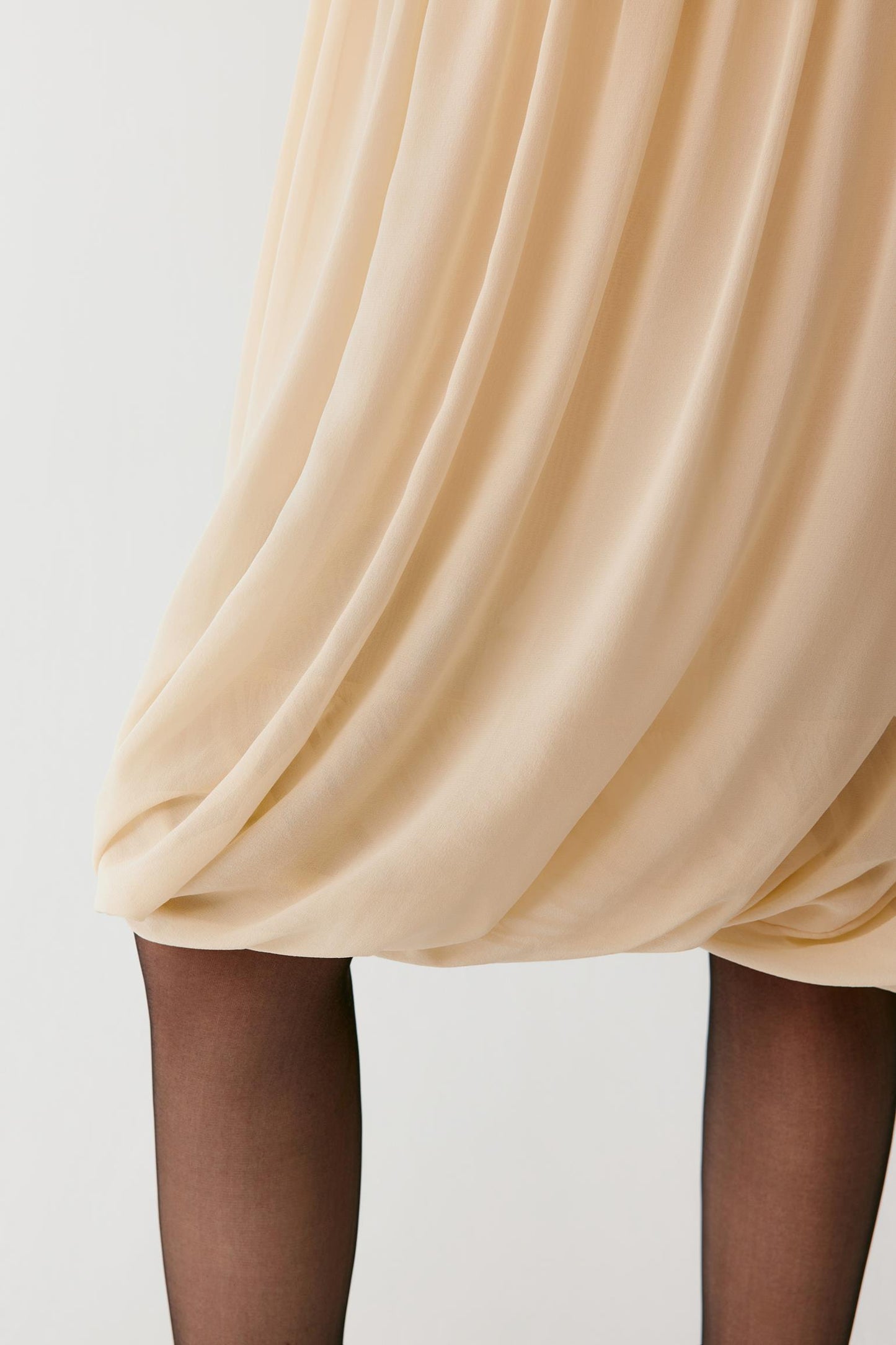Close-up of a draped, pleated beige skirt with sheer black tights worn by a model, set against a plain light gray background.