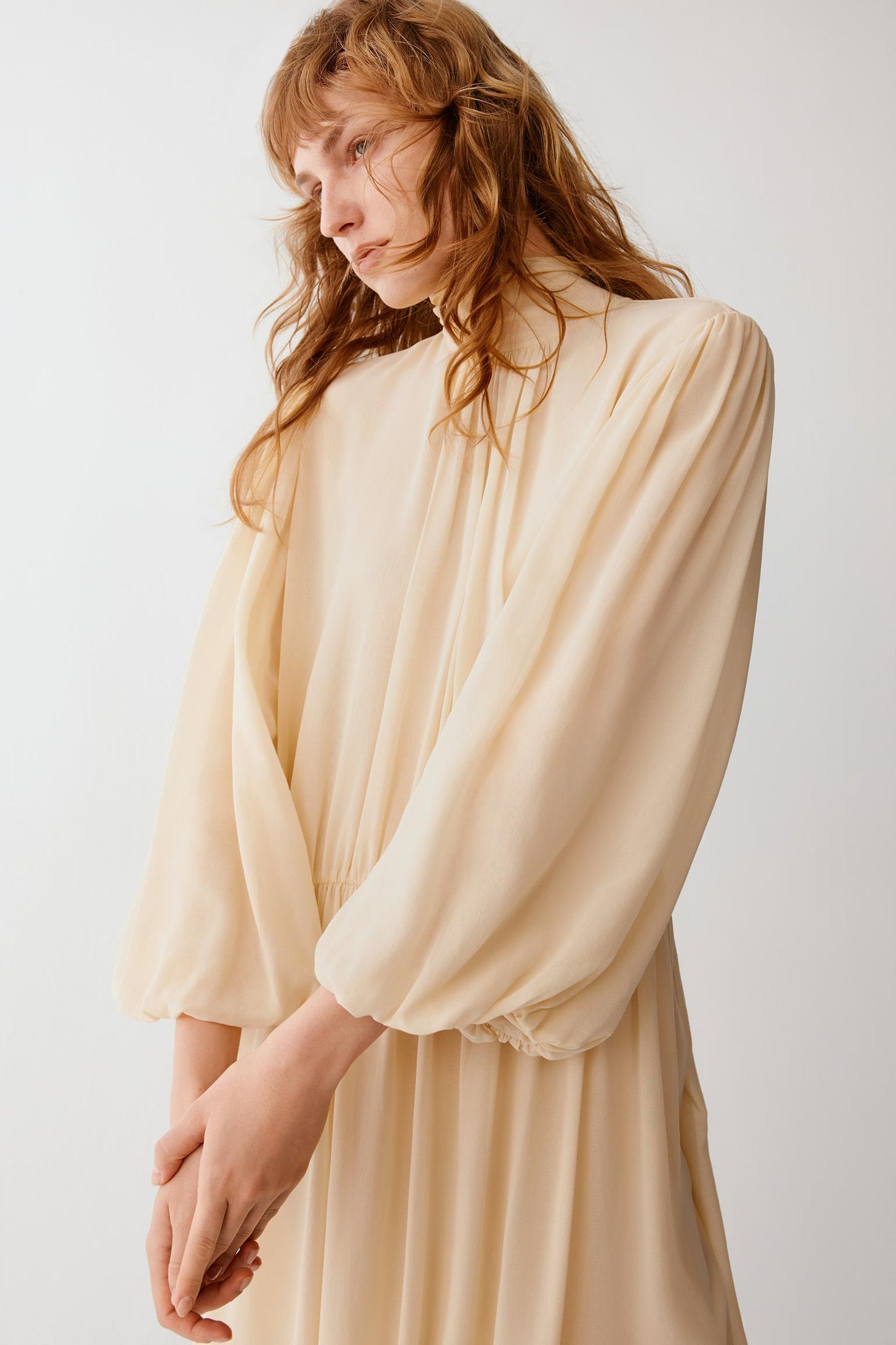 A woman wearing a cream-colored, high-neck dress with voluminous balloon sleeves and a gathered waist, photographed against a plain light gray background.