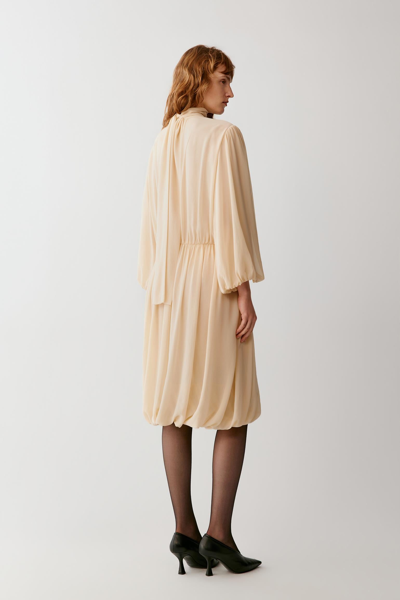 A woman shown from the back wearing a light beige midi dress with three-quarter sleeves, black tights, and black heels, photographed against a plain light gray studio background.