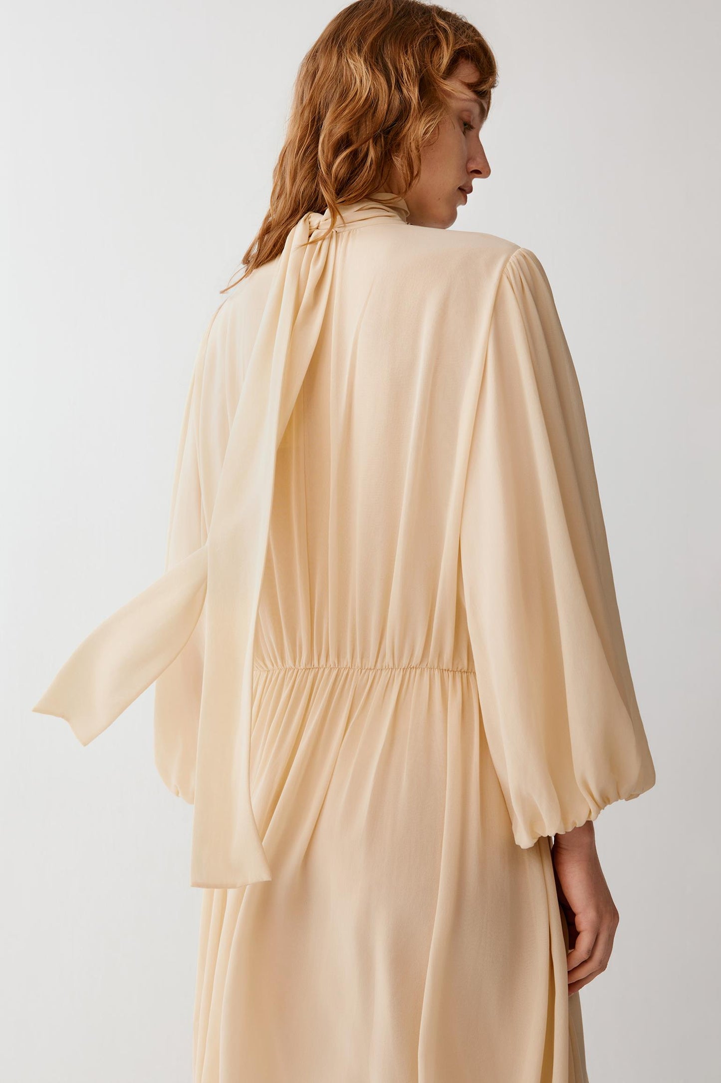 A woman wearing a flowing, light peach-beige long-sleeve dress, shown from the back against a plain light gray studio background.