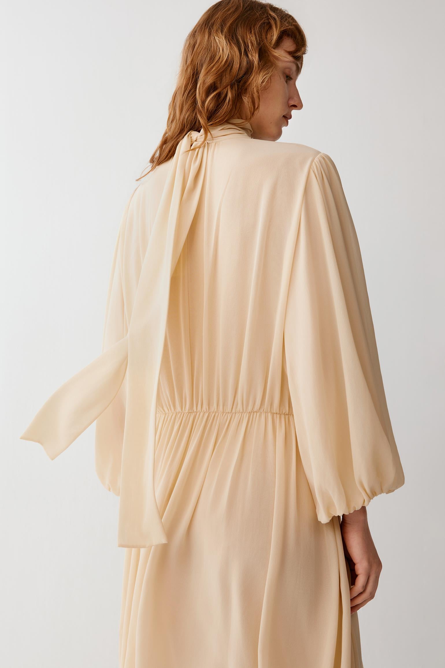 A woman wearing a flowing, light peach-beige long-sleeve dress, shown from the back against a plain light gray studio background.