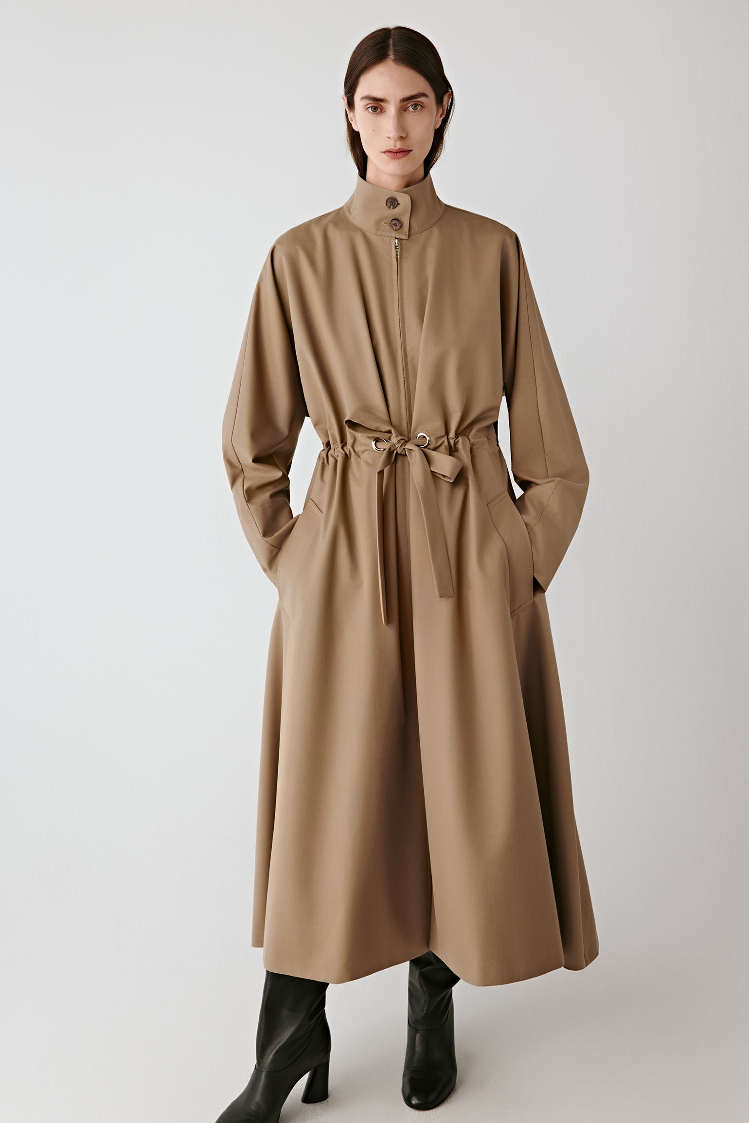Front view of a long light beige coat with a tied belt at the waist, straight cut, worn with black boots.