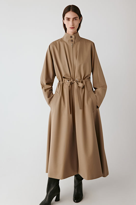 Front view of a long light beige coat with a tied belt at the waist, straight cut, worn with black boots.