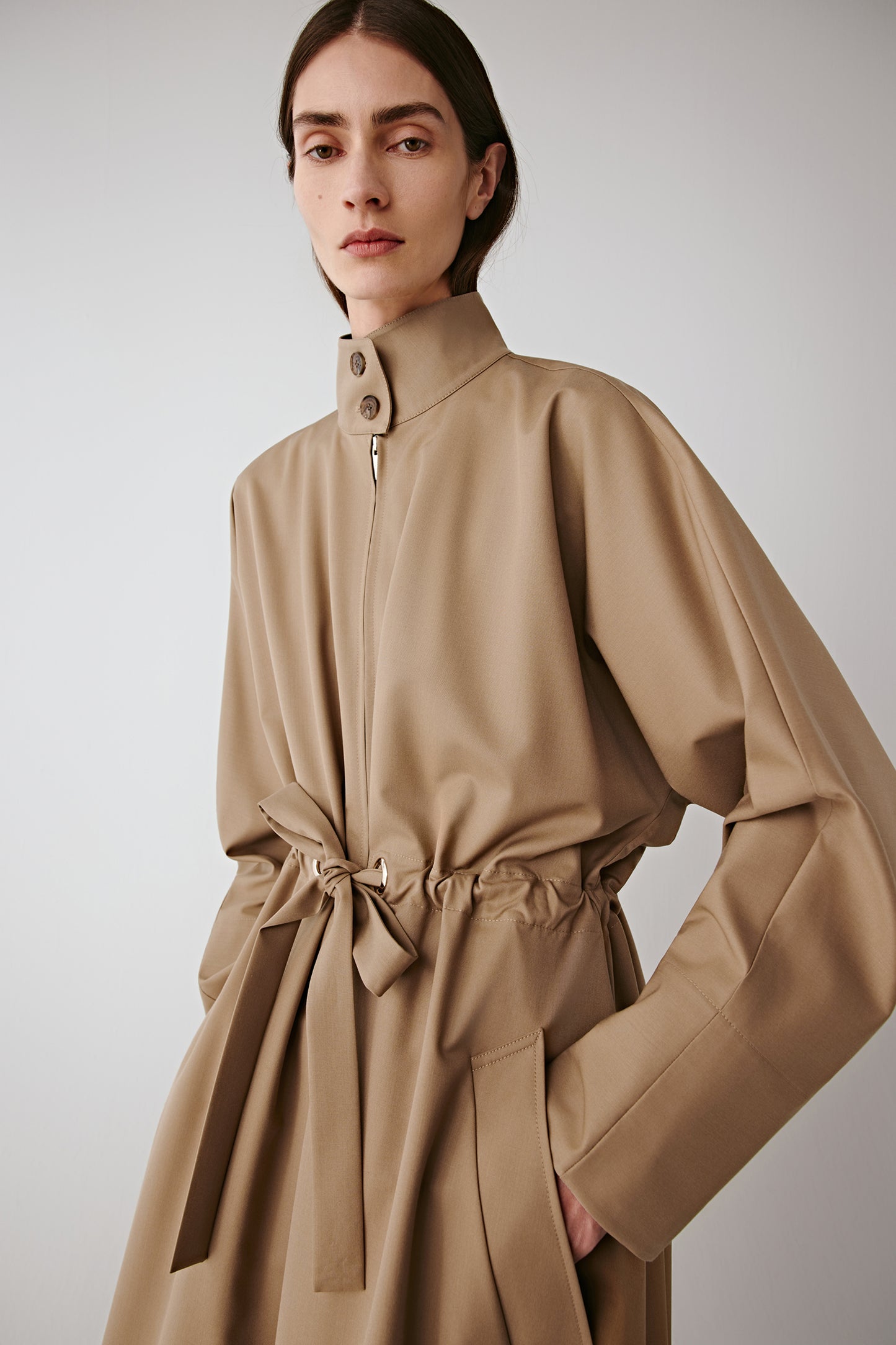 Side view of the beige coat with hands in pockets, highlighting the length and tailored fit.